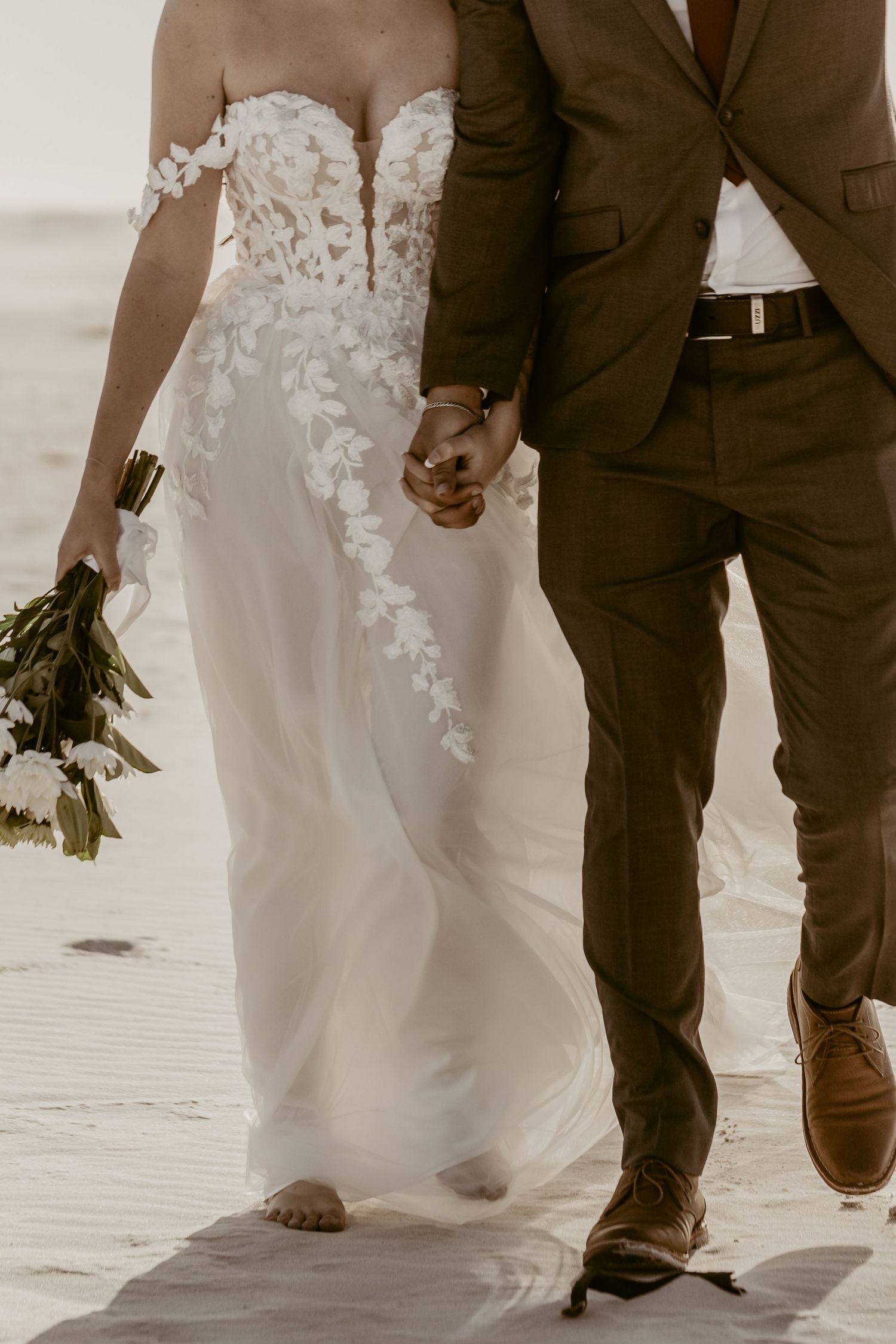Close-up of bride in lace floral wedding gown holding bouquet, walking with groom in brown suit on beach.