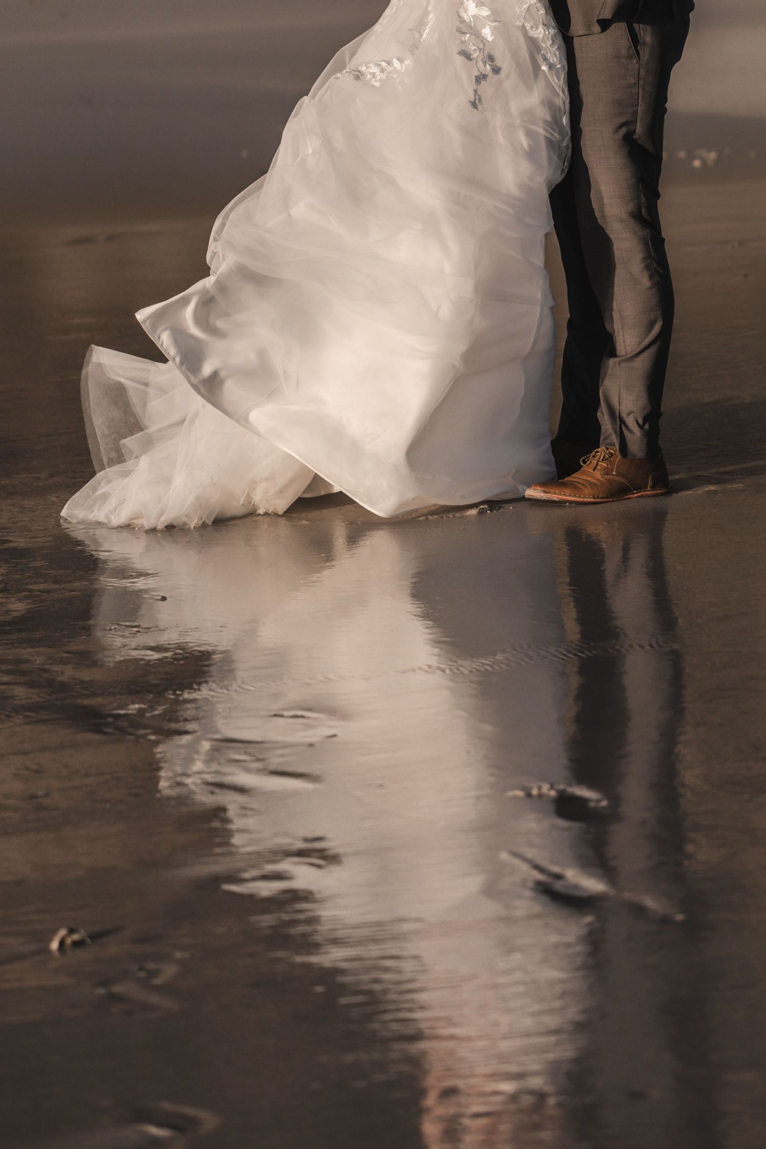 Bride in flowing white wedding gown and groom in gray pants standing on reflective wet beach sand.