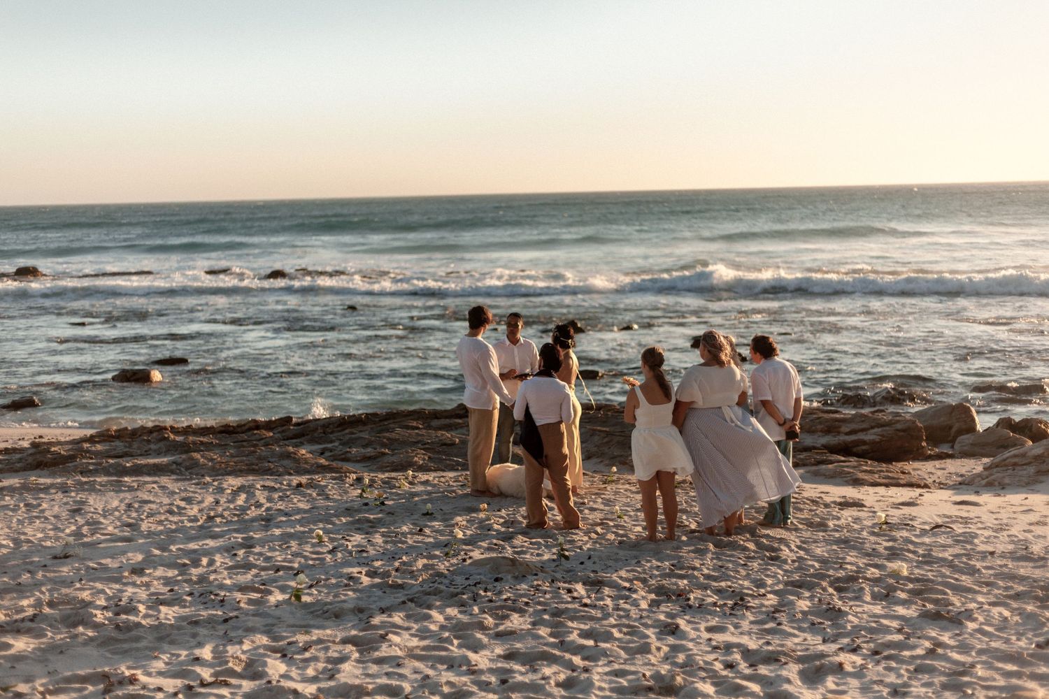 A group in white attire gathered on a beach at sunset watching waves crash against rocky shores.