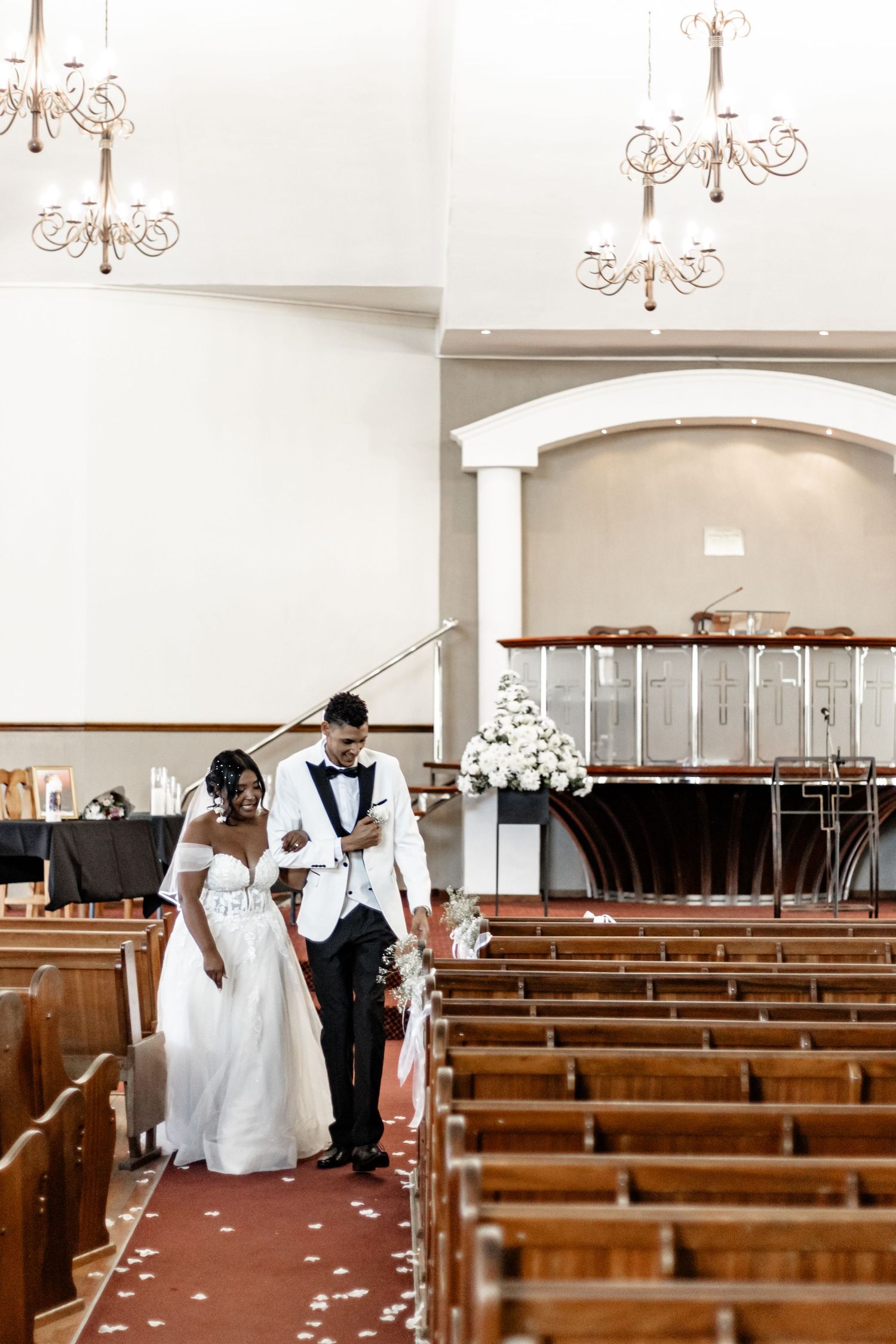 Wedding party members walk down the aisle of a church adorned with chandeliers and wooden pews.