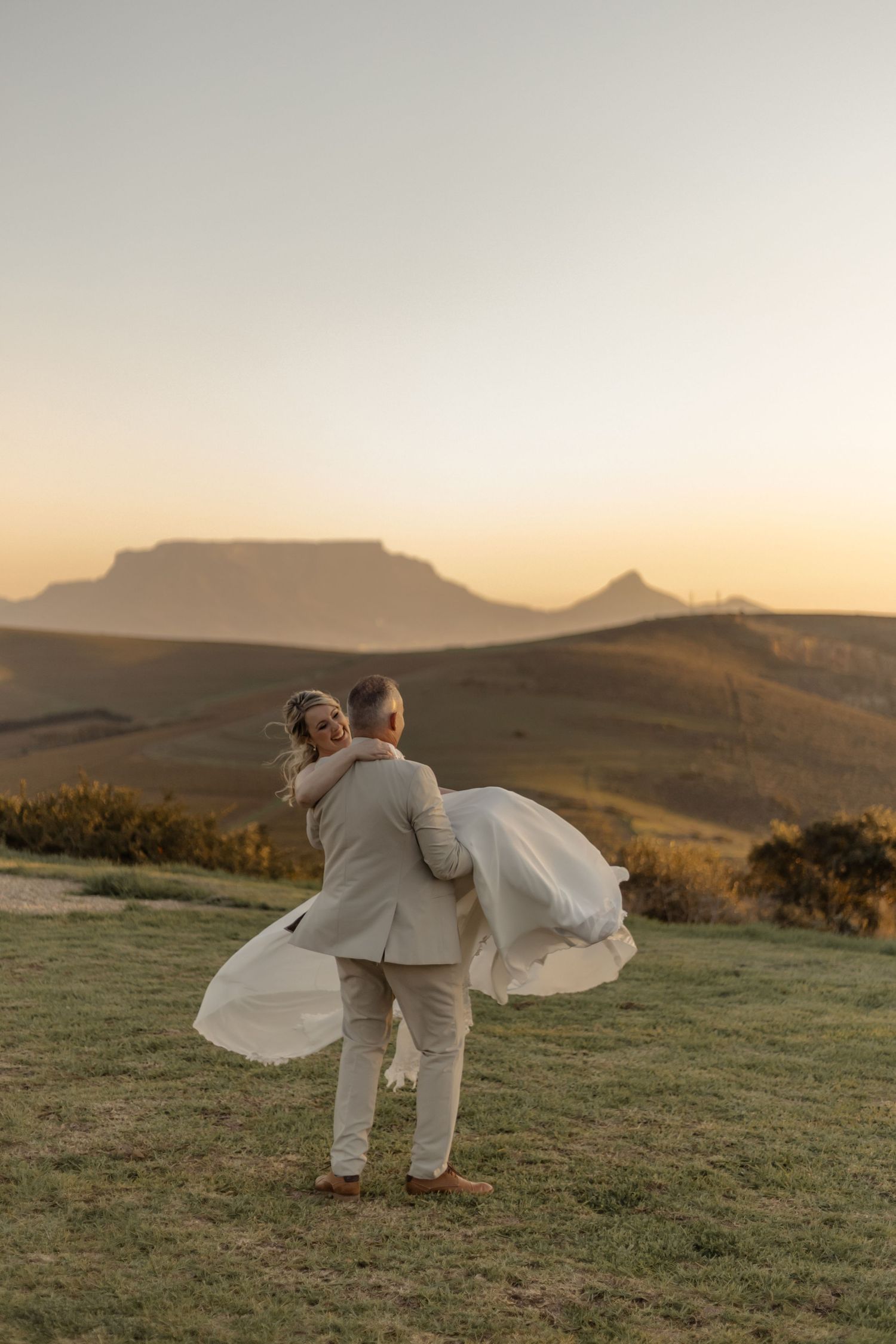 A couple embraces in matching white outfits on a grassy hillside during golden hour with mountains in the background.