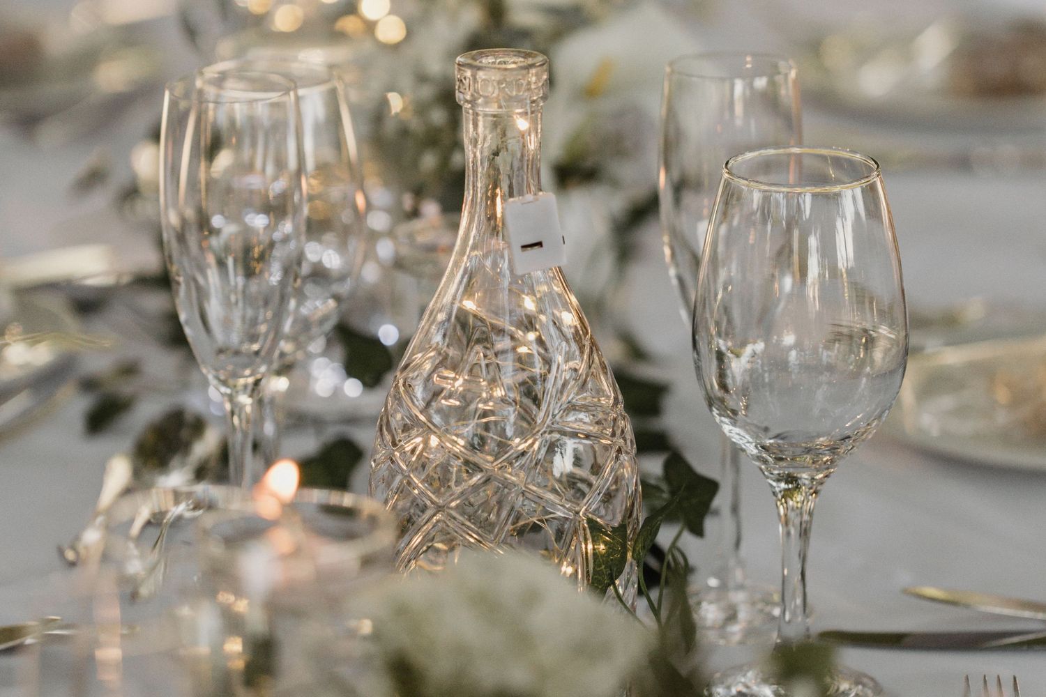 Elegant wine glasses and a decorative crystal bottle on a table with soft bokeh lights and greenery in the background.