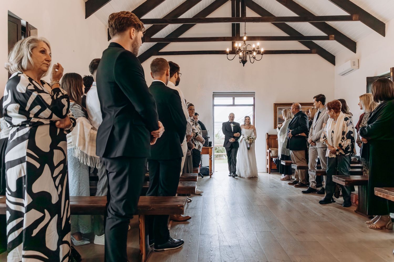 Bride walks down the aisle in a white chapel wedding ceremony with guests standing in wooden pews.