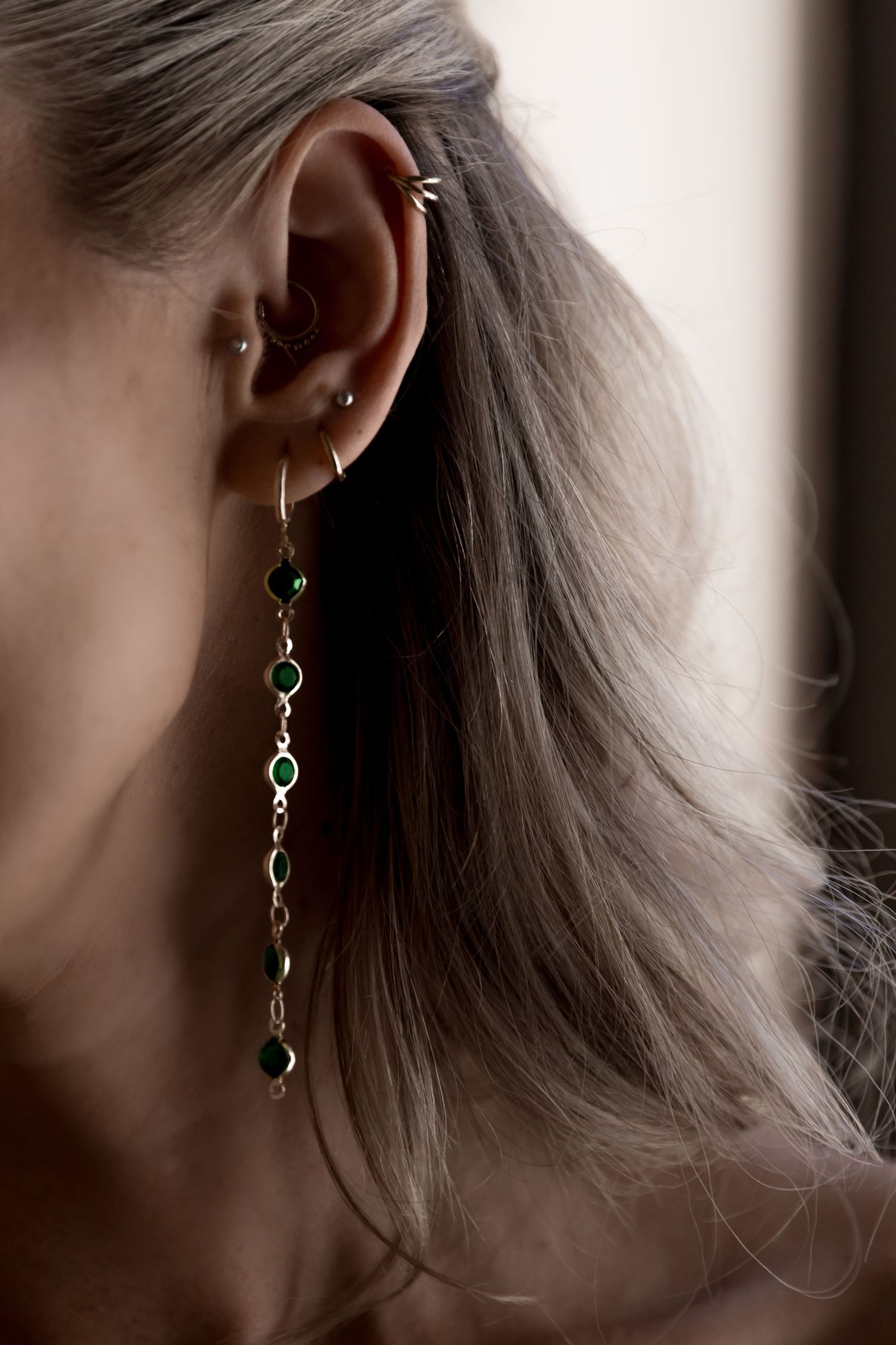 Close-up of a delicate dangling earring with green gemstones against soft gray hair in natural lighting.