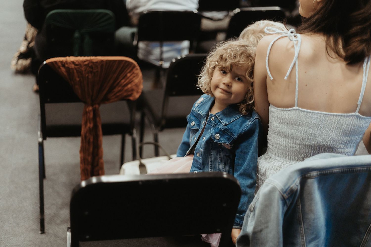 A woman in a denim jacket and white dress holds a young child while sitting outdoors.