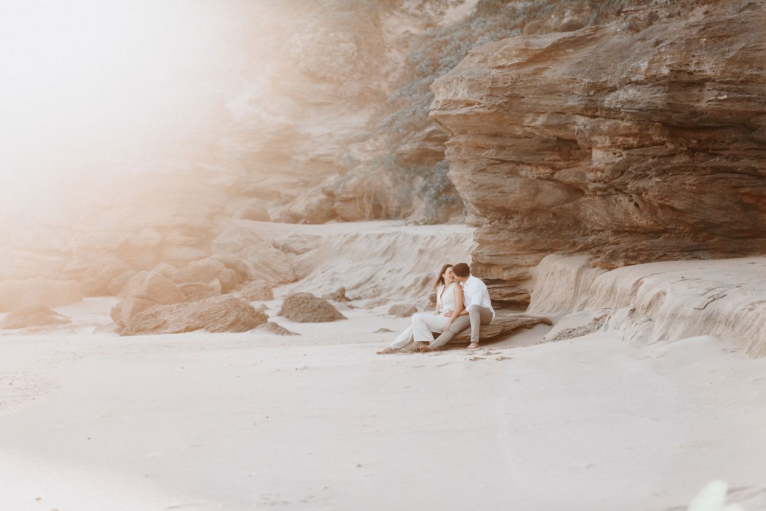 Two people sitting together on a rocky beach at sunset with dramatic cliffs in the background.