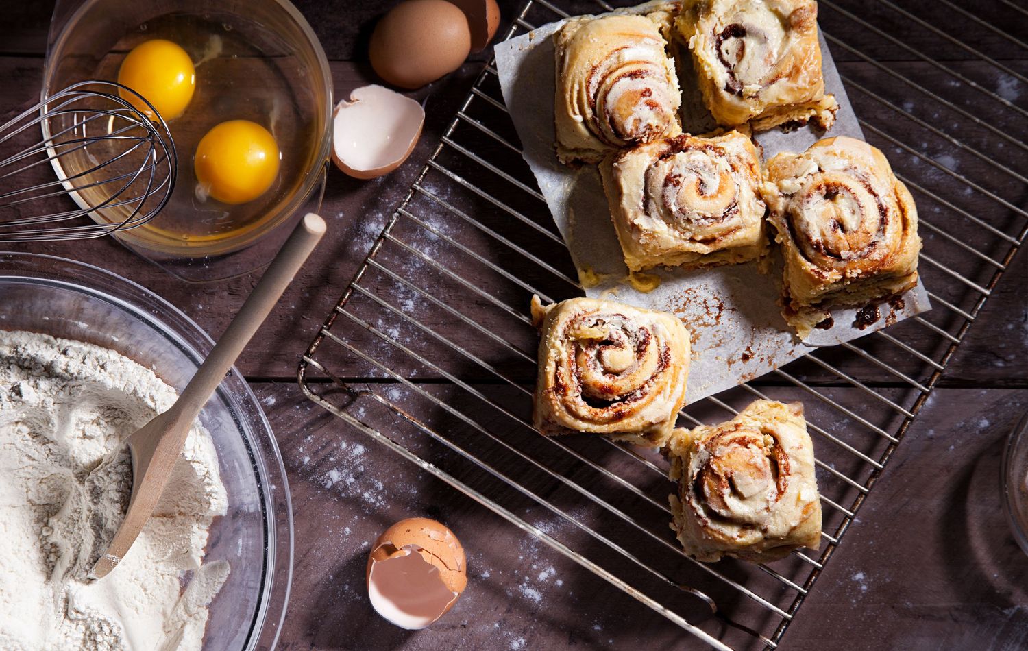 Raw eggs and cinnamon roll pastries cooling on a wire rack with baking ingredients in a dark rustic setting.