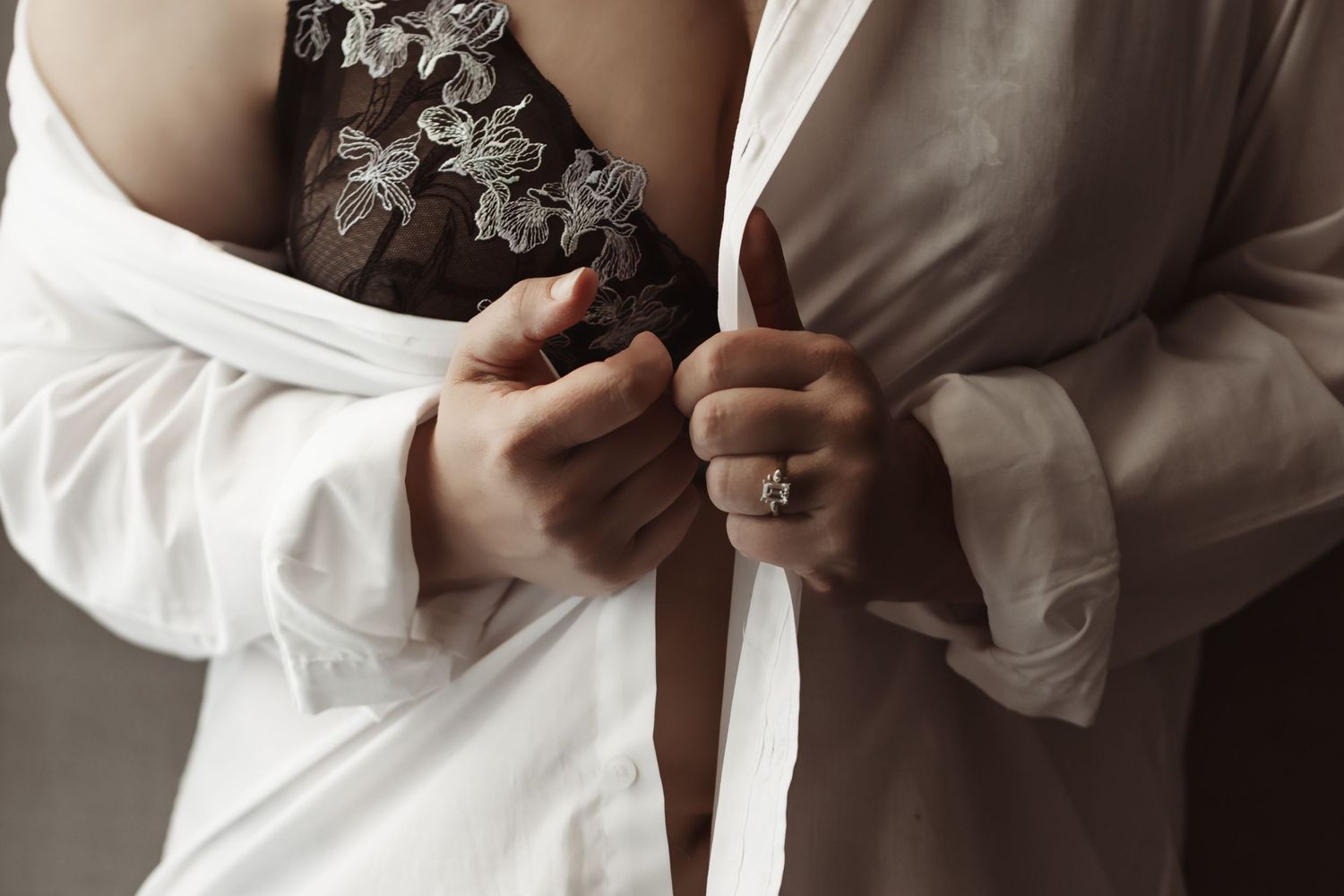 Close-up of hands buttoning a white dress shirt with delicate lace detail and a sparkling engagement ring.