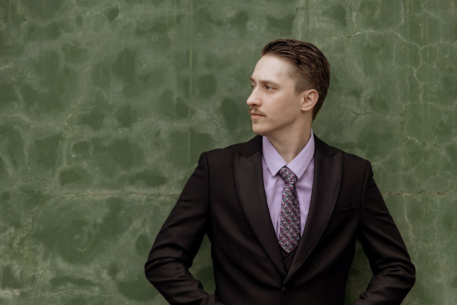 Professional headshot photo of someone in a black suit and purple tie against a textured green wall background.