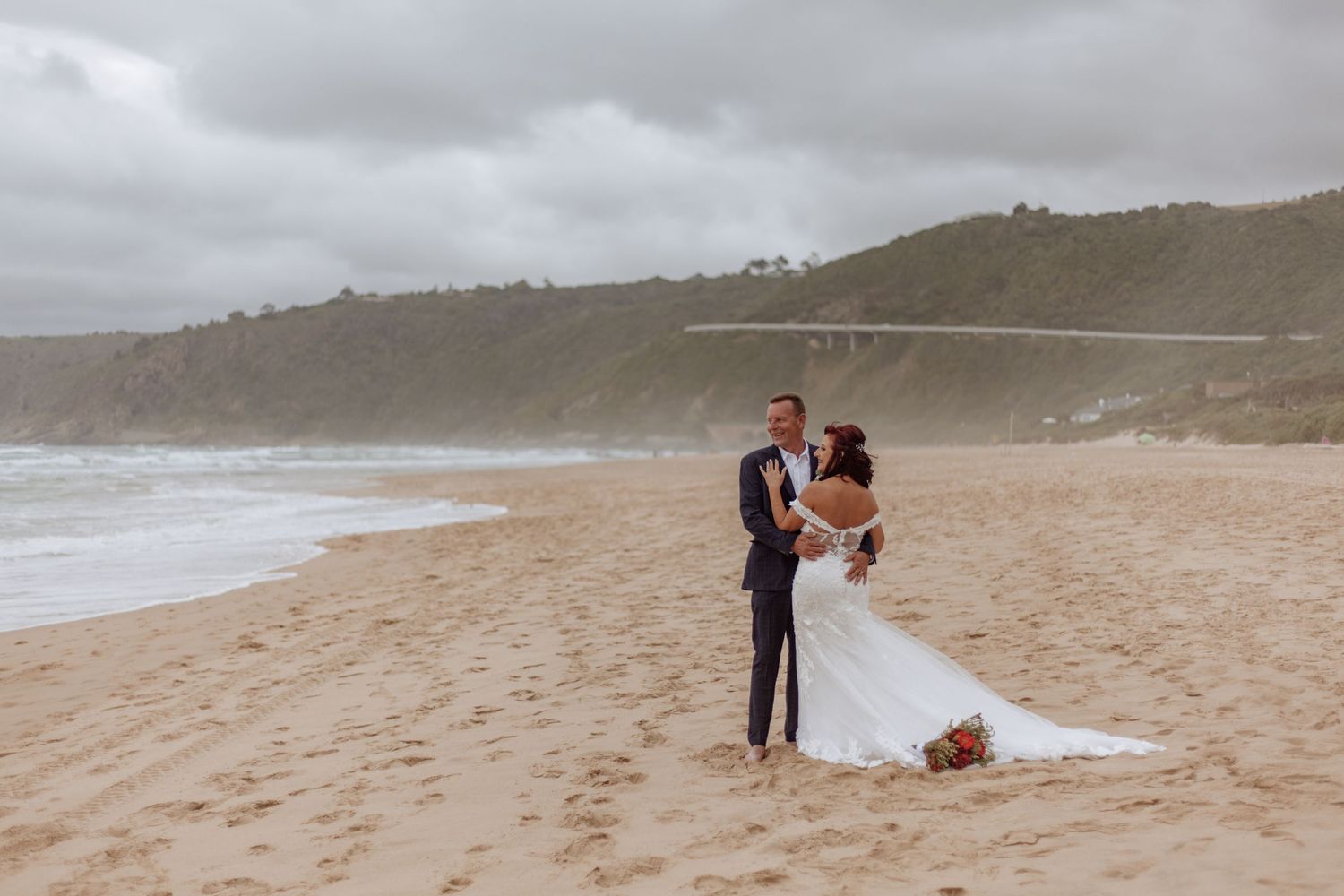 A couple in wedding attire embraces on a misty beach with cliffs in the background.
