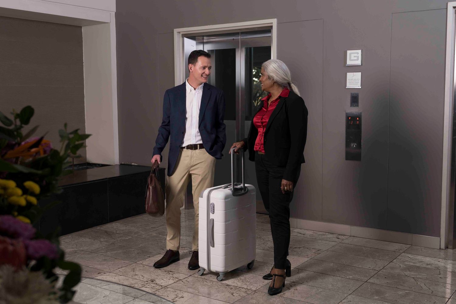 Two people chatting in a lobby area next to luggage and decorative plants.