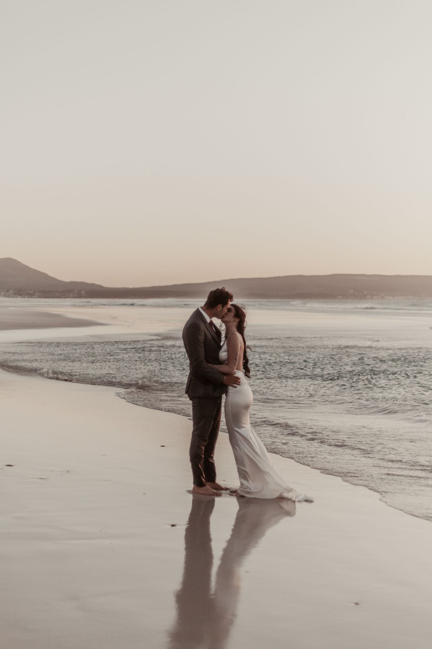 Bride and groom sharing a romantic kiss on a serene beach at sunset, waves gently lapping at their feet.