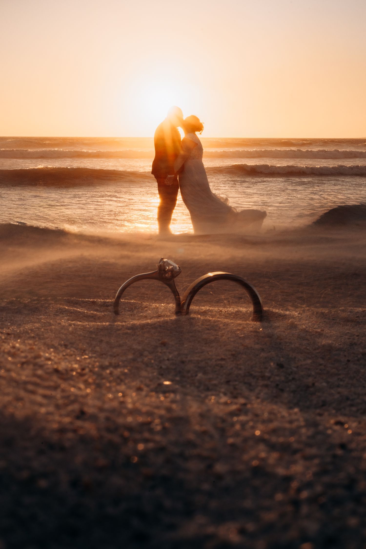 Couple kissing at sunset on beach with wedding rings in foreground sand, romantic golden hour ocean backdrop.