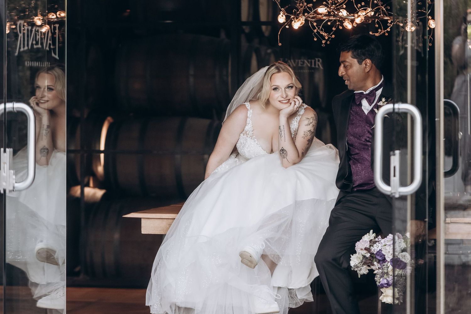 Bride in white gown and groom in purple bow tie pose elegantly at a winery entrance with barrel decor.