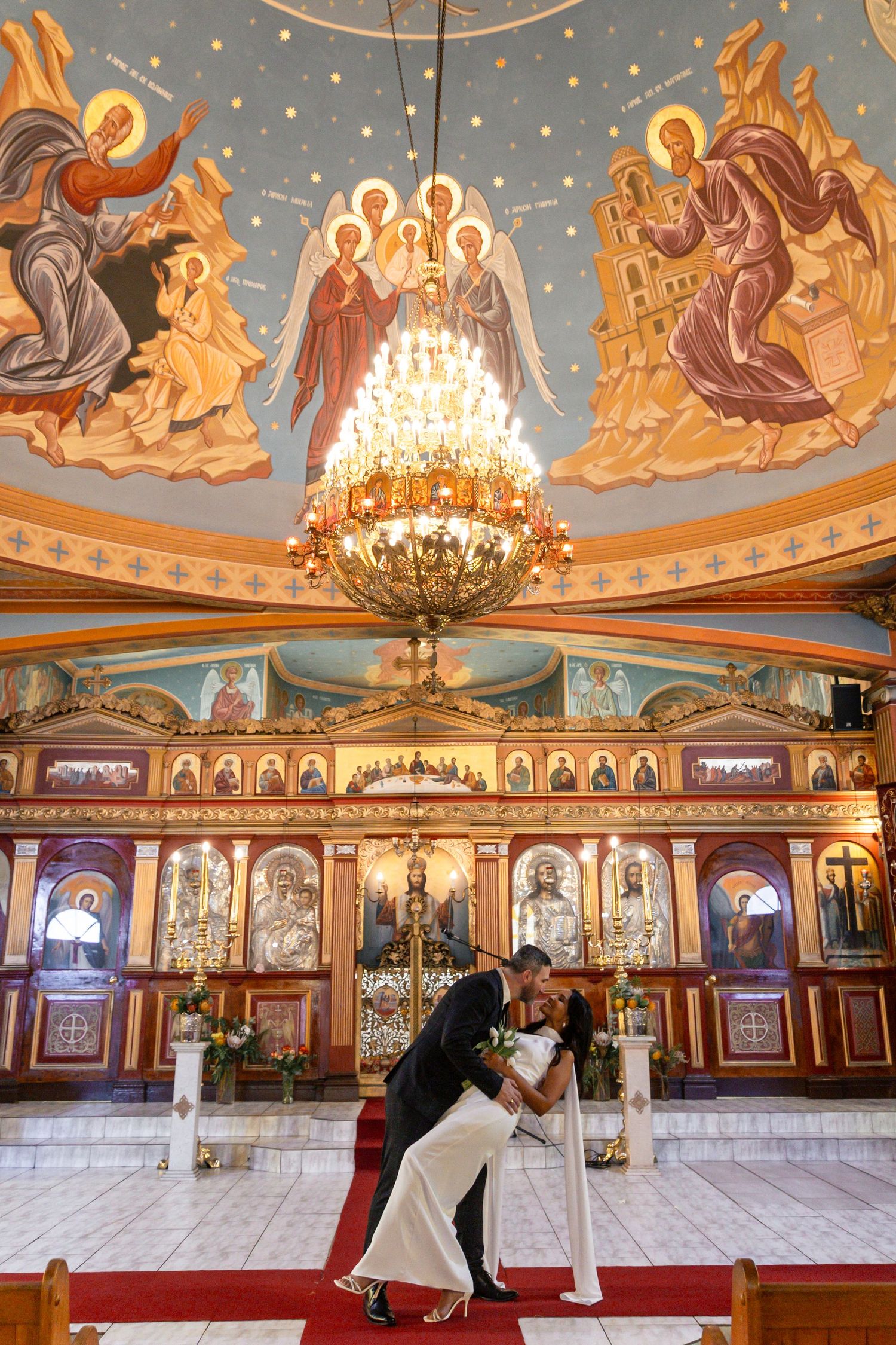 Couple sharing a romantic dip kiss inside an ornate Orthodox church with Byzantine frescoes and grand chandelier.