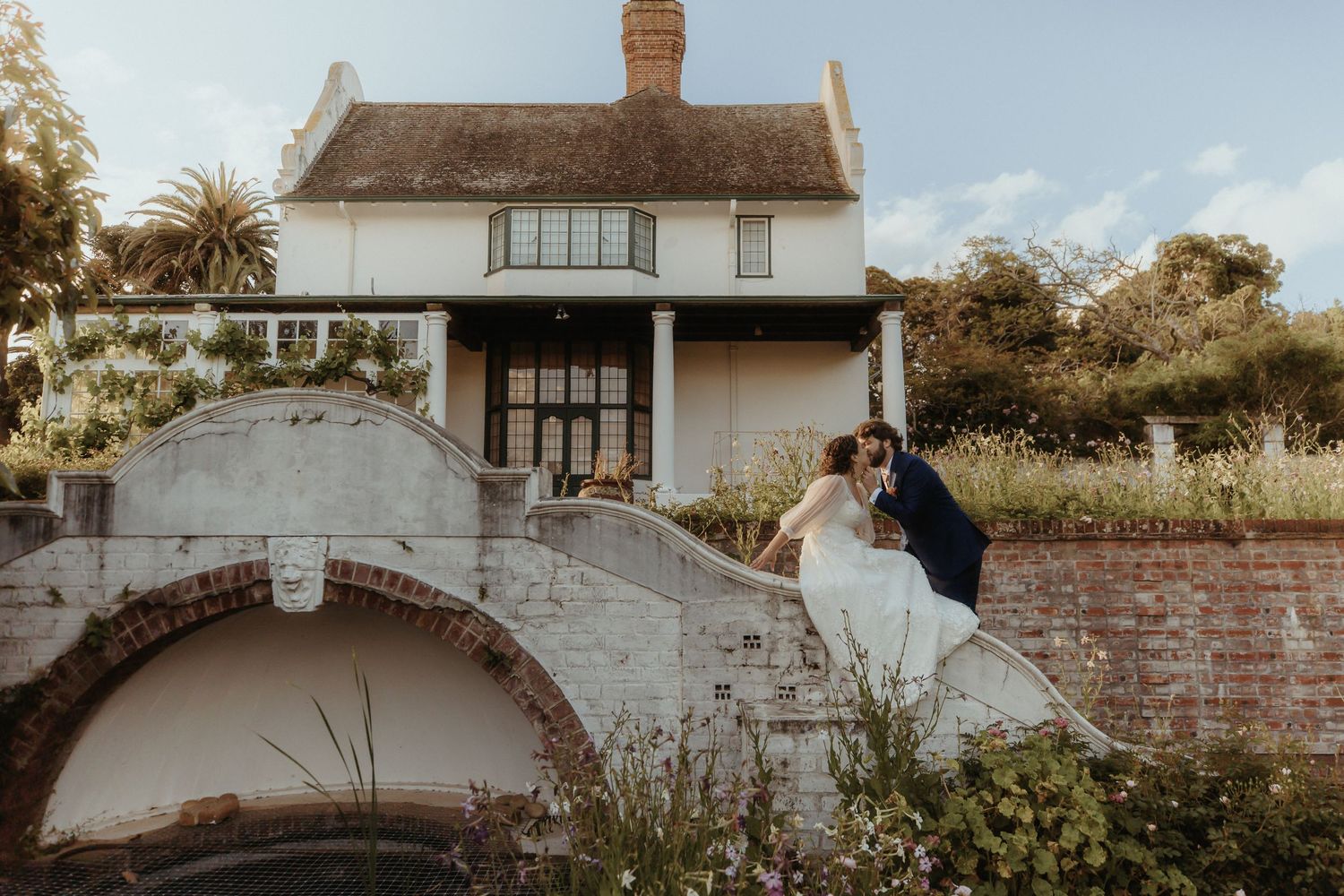 A couple shares a romantic kiss on a stone bridge outside a charming white country house surrounded by lush gardens.