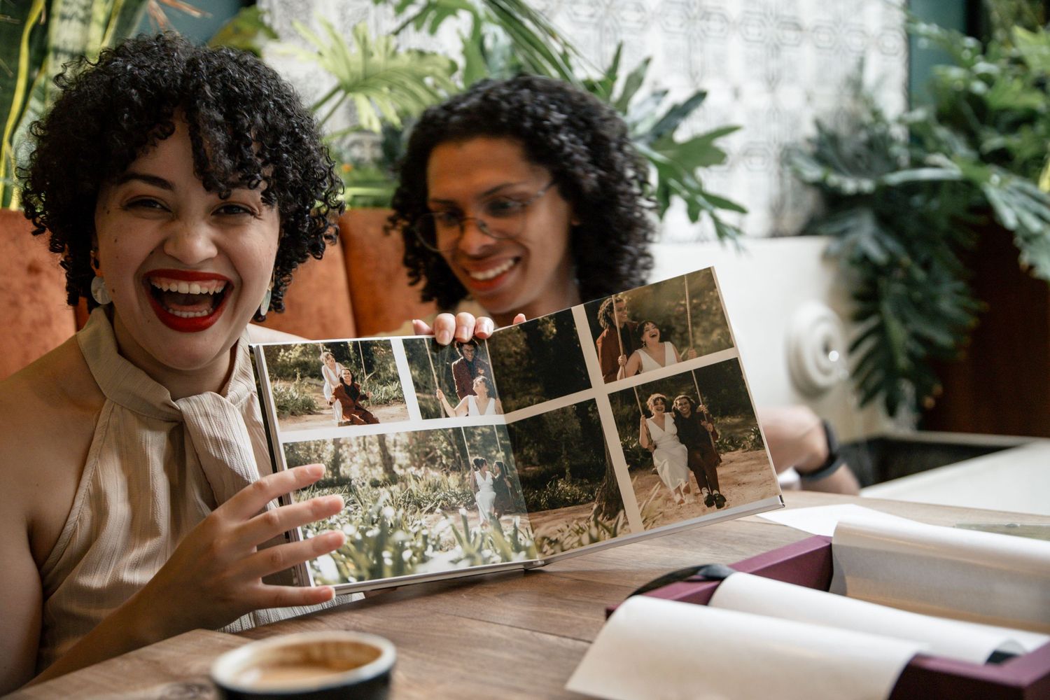 Two women smiling and laughing while looking at a wedding photo album together at a cafe table.