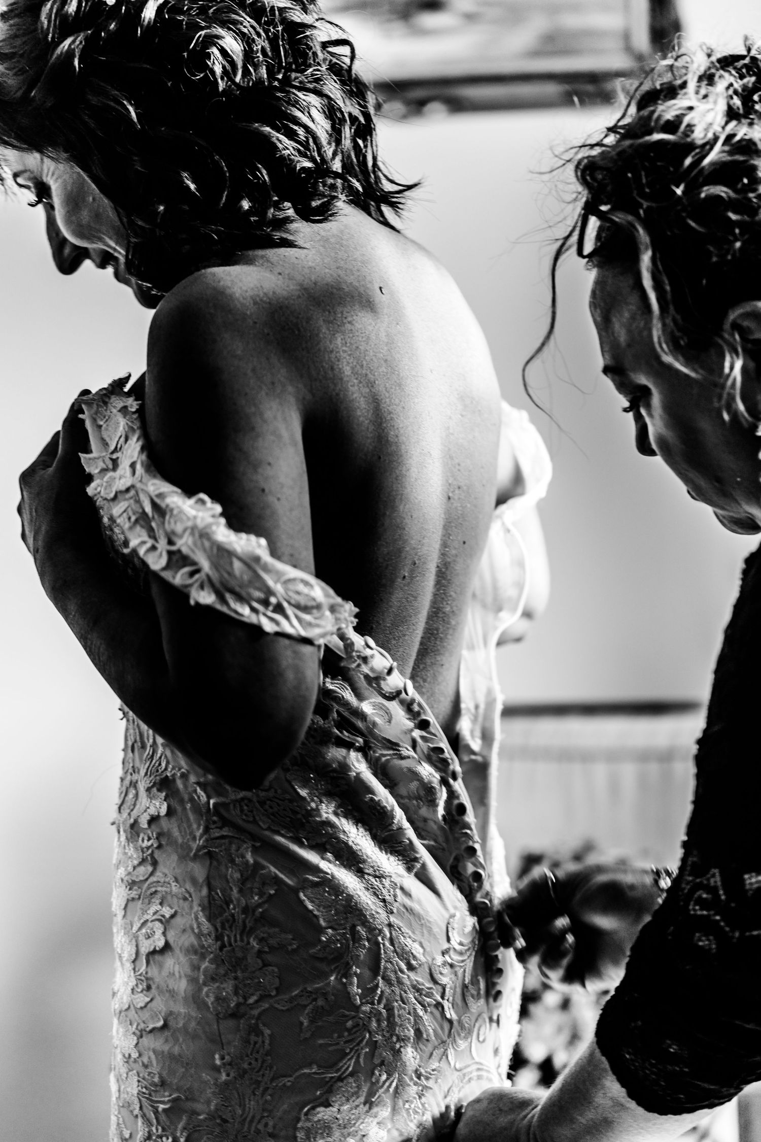 A black and white photograph of someone in a lace dress being helped with dress adjustments from behind.