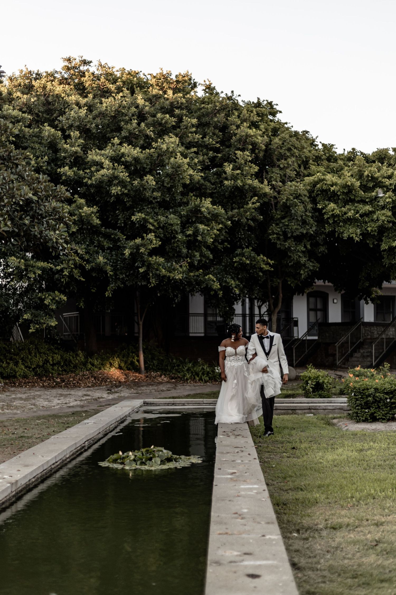 A couple in wedding attire walking along a reflecting pool with lush greenery in the background.
