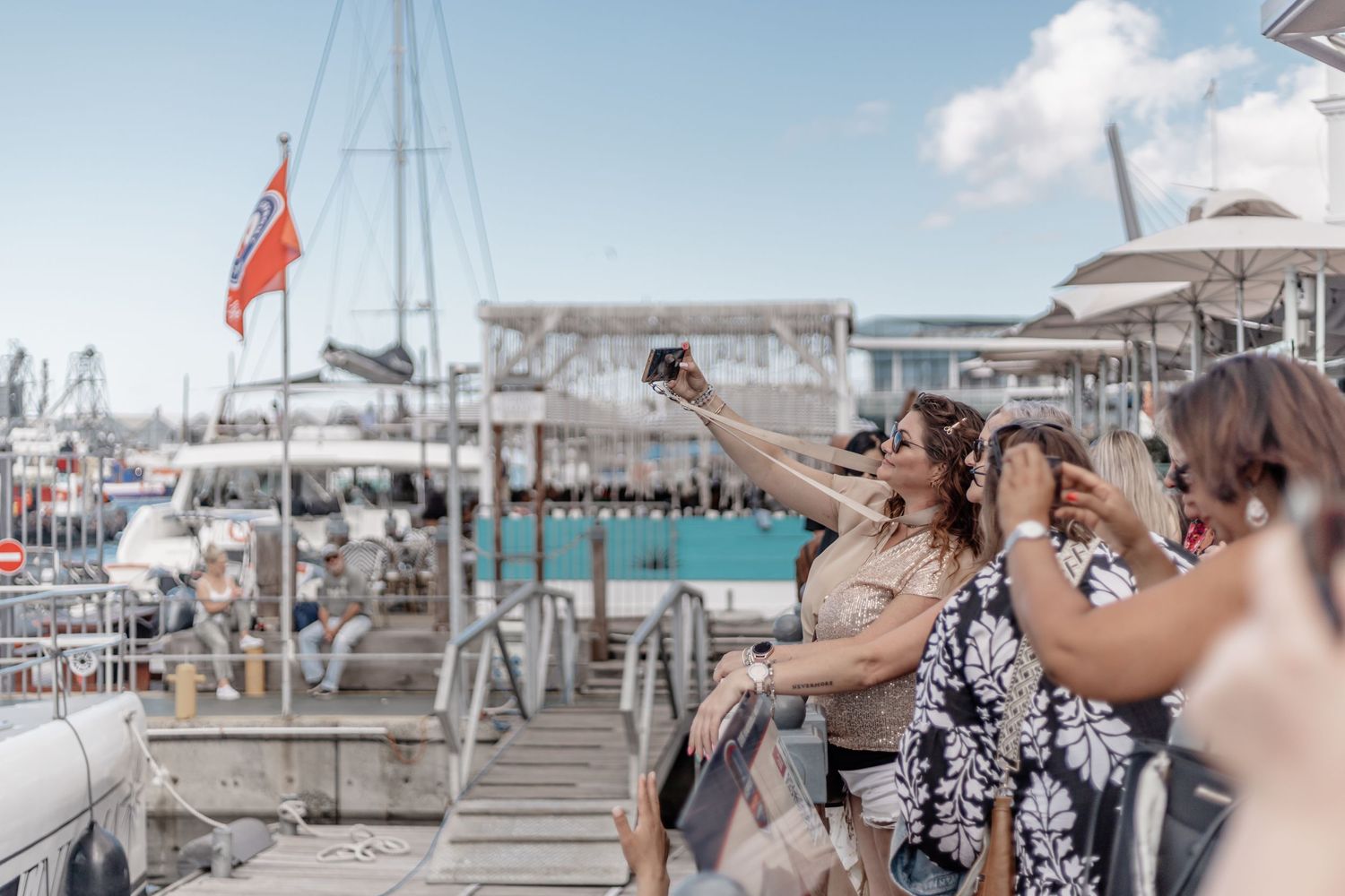 Tourists taking photos from a yacht or boat deck with turquoise water and boats in the background.