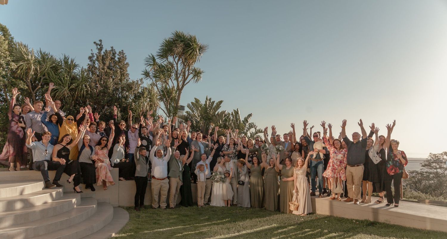 Large group of wedding guests celebrating with raised arms at an outdoor venue during sunset.