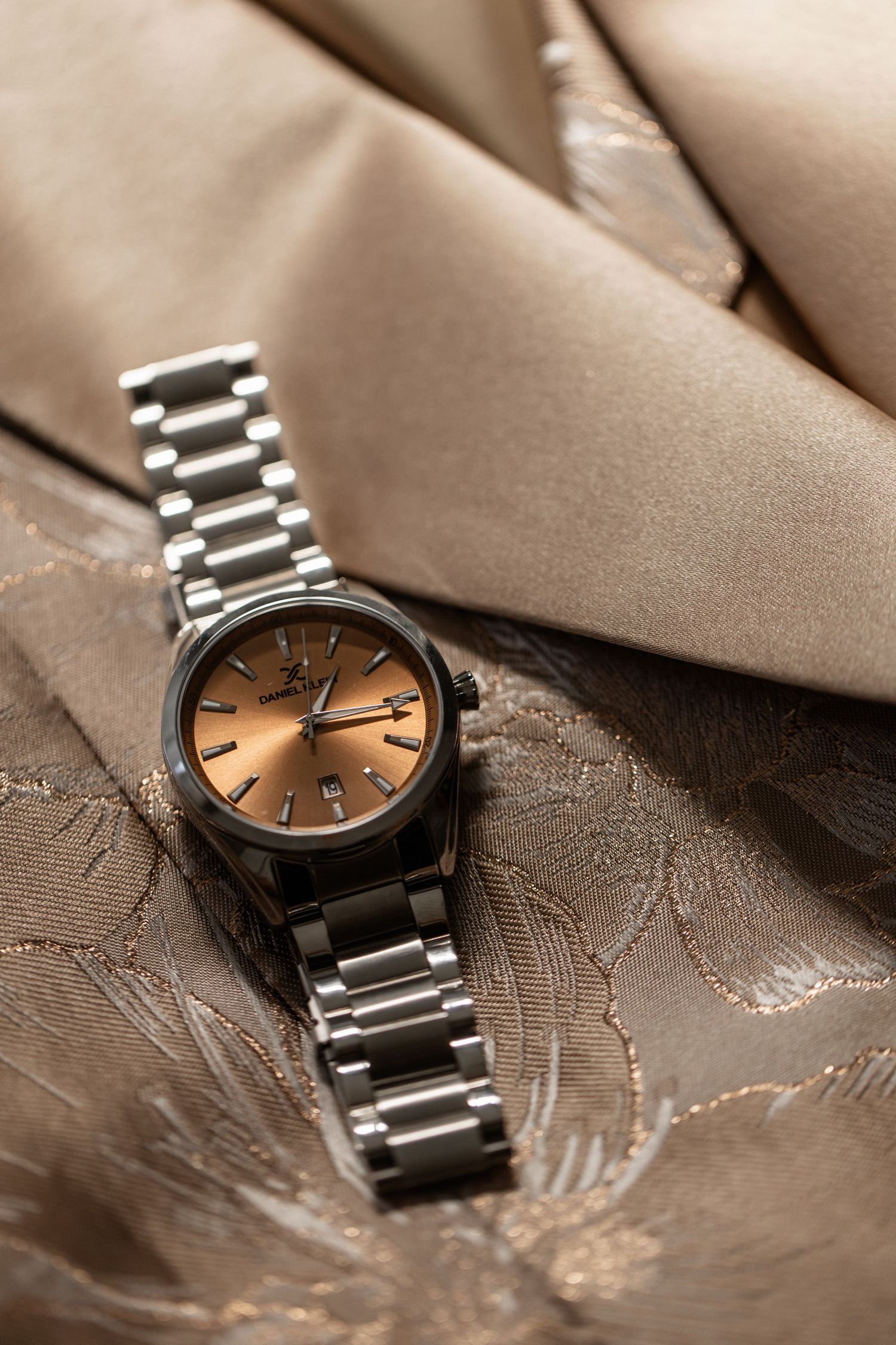Elegant silver wristwatch with copper-toned face lying on textured brown satin fabric.