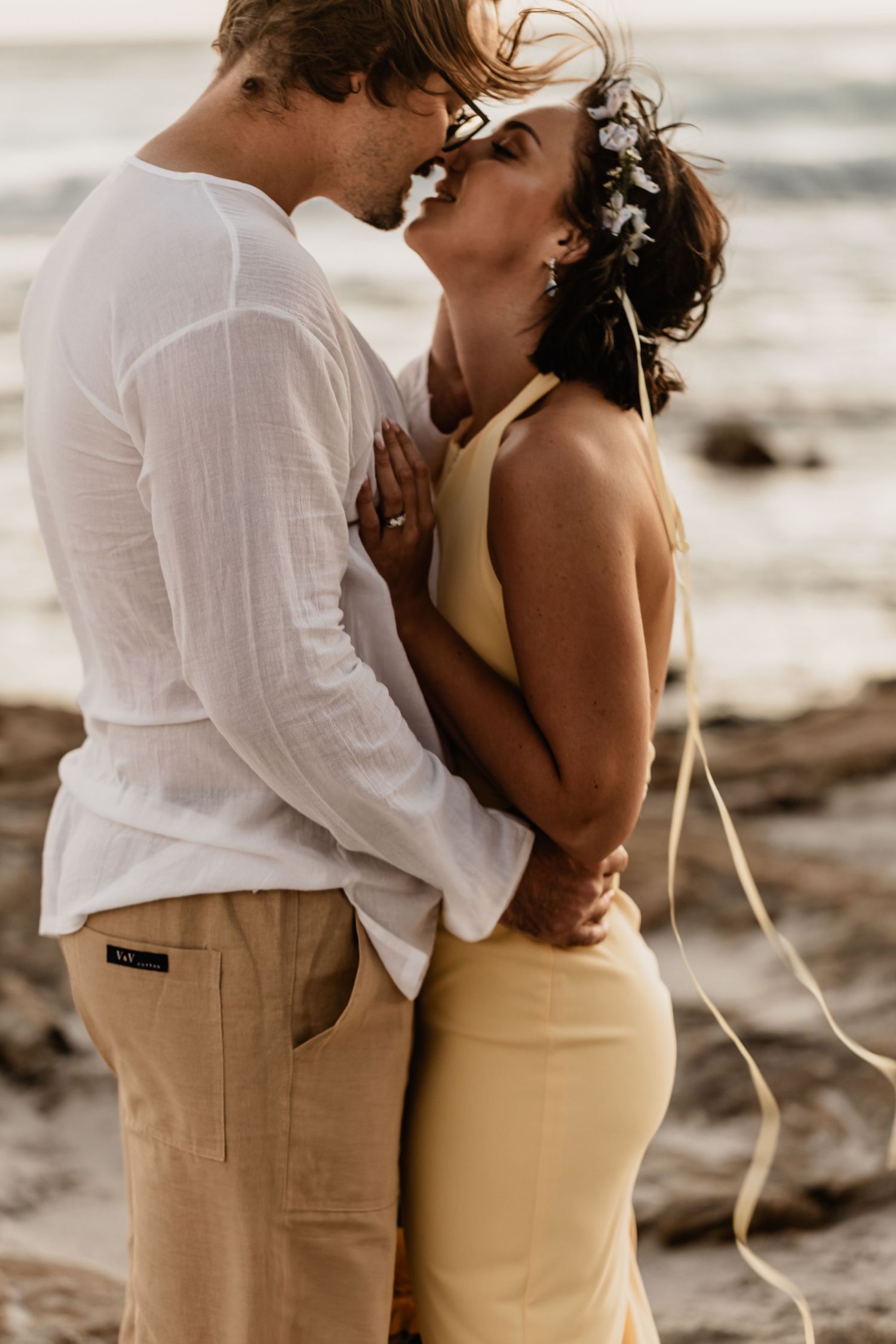 A romantic couple embraces on a sandy beach at sunset, wearing white and beige outfits.