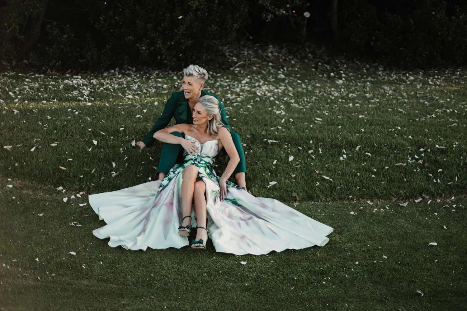 A couple sits together on grass surrounded by scattered white flower petals during a romantic outdoor photoshoot.