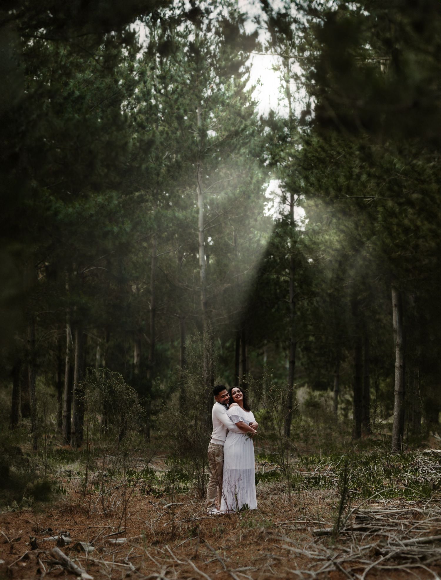 A couple in white outfits embraces in a misty forest setting with tall trees creating a romantic atmosphere.