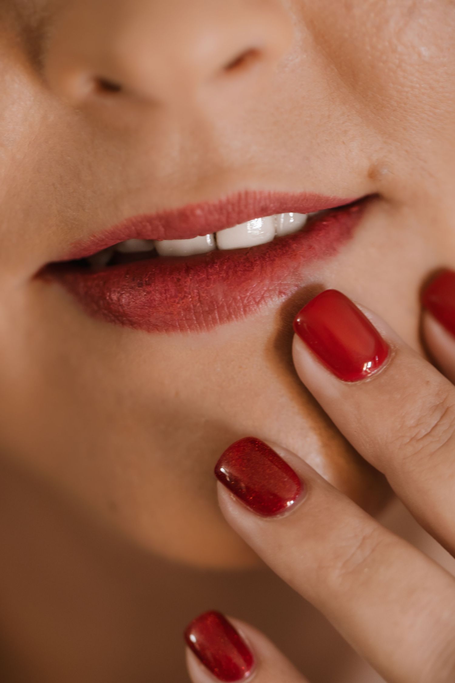 Close-up of red lipstick and matching red nail polish shown against neutral beige background.