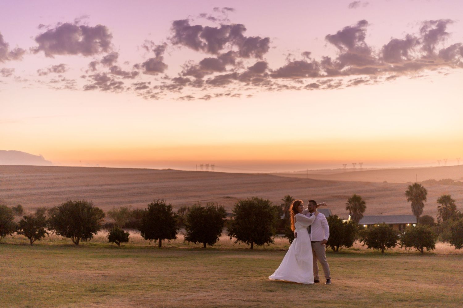 A romantic wedding couple embraces in a golden field at sunset with dramatic clouds and trees in the background.