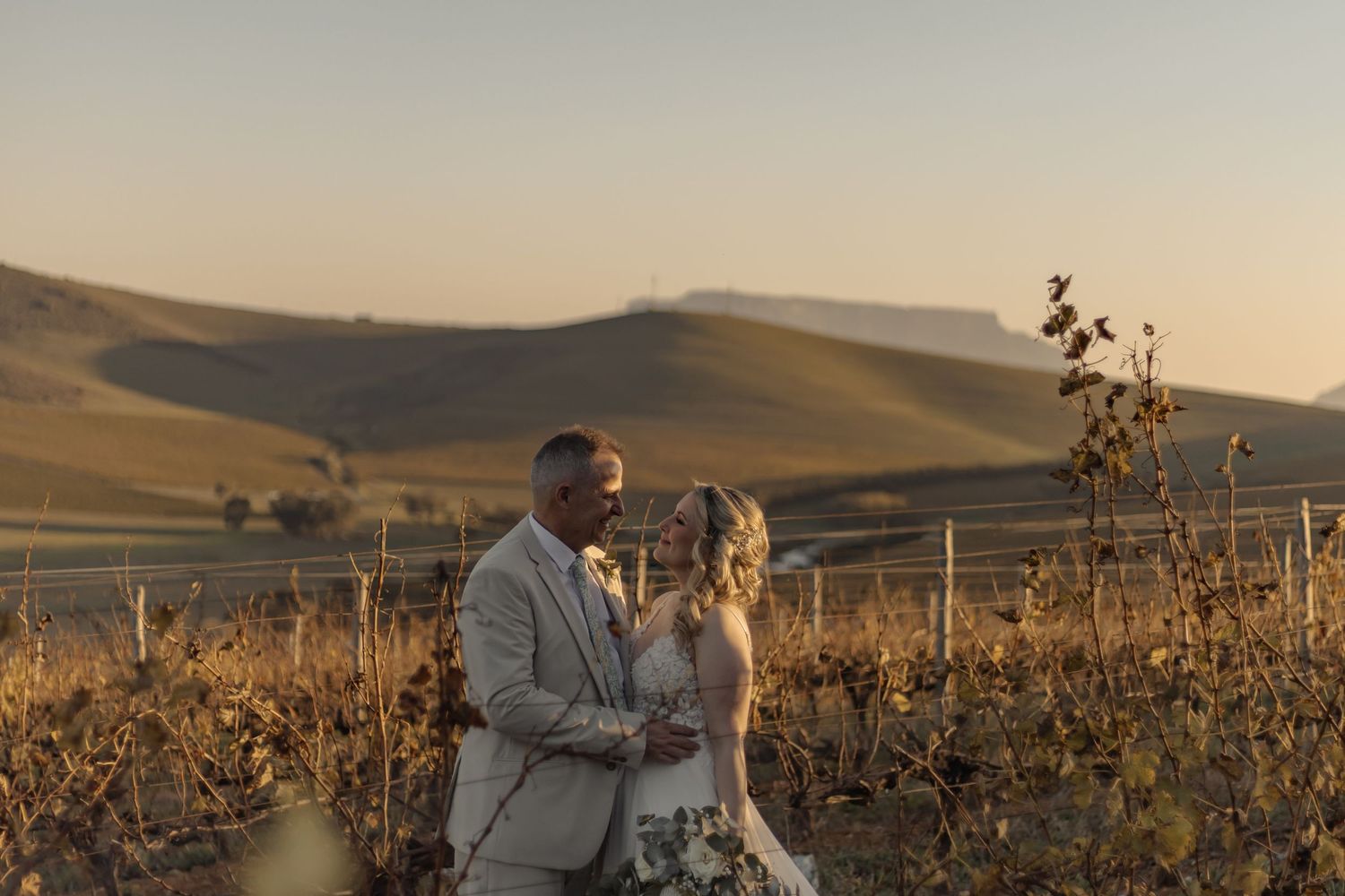 A couple shares an intimate embrace among vineyard rows at sunset in rolling hills countryside.