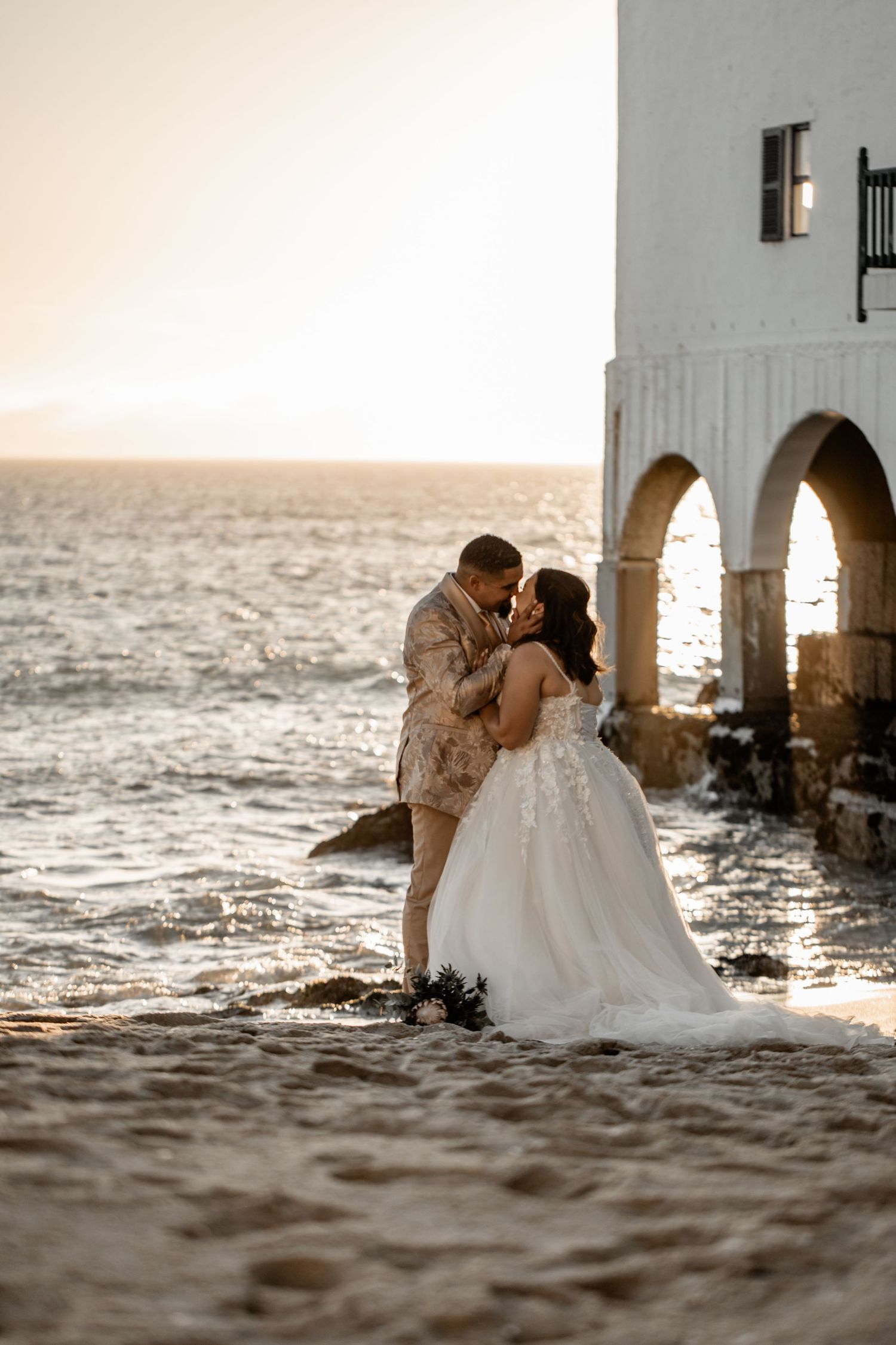 A couple shares a romantic kiss on the beach at sunset with water and arched pillars in the background.