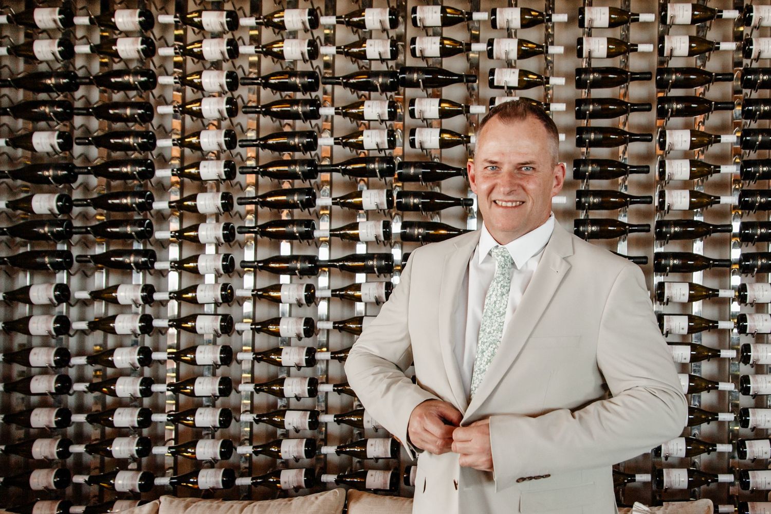 A person in a light beige suit and tie stands in front of an extensive wall-mounted wine bottle collection.