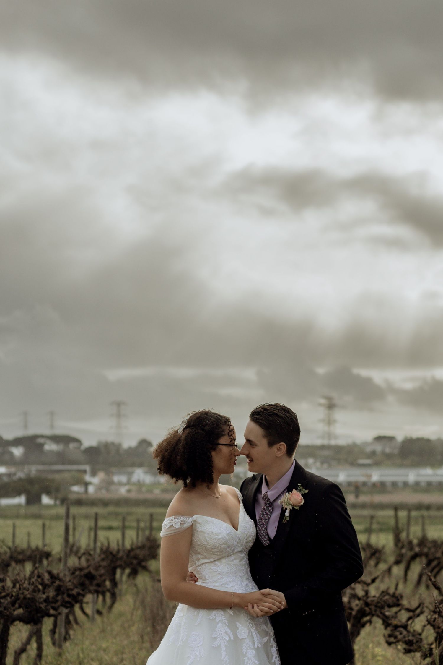 A couple embraces in a cloudy vineyard setting with dramatic stormy skies overhead during their romantic wedding photo session.
