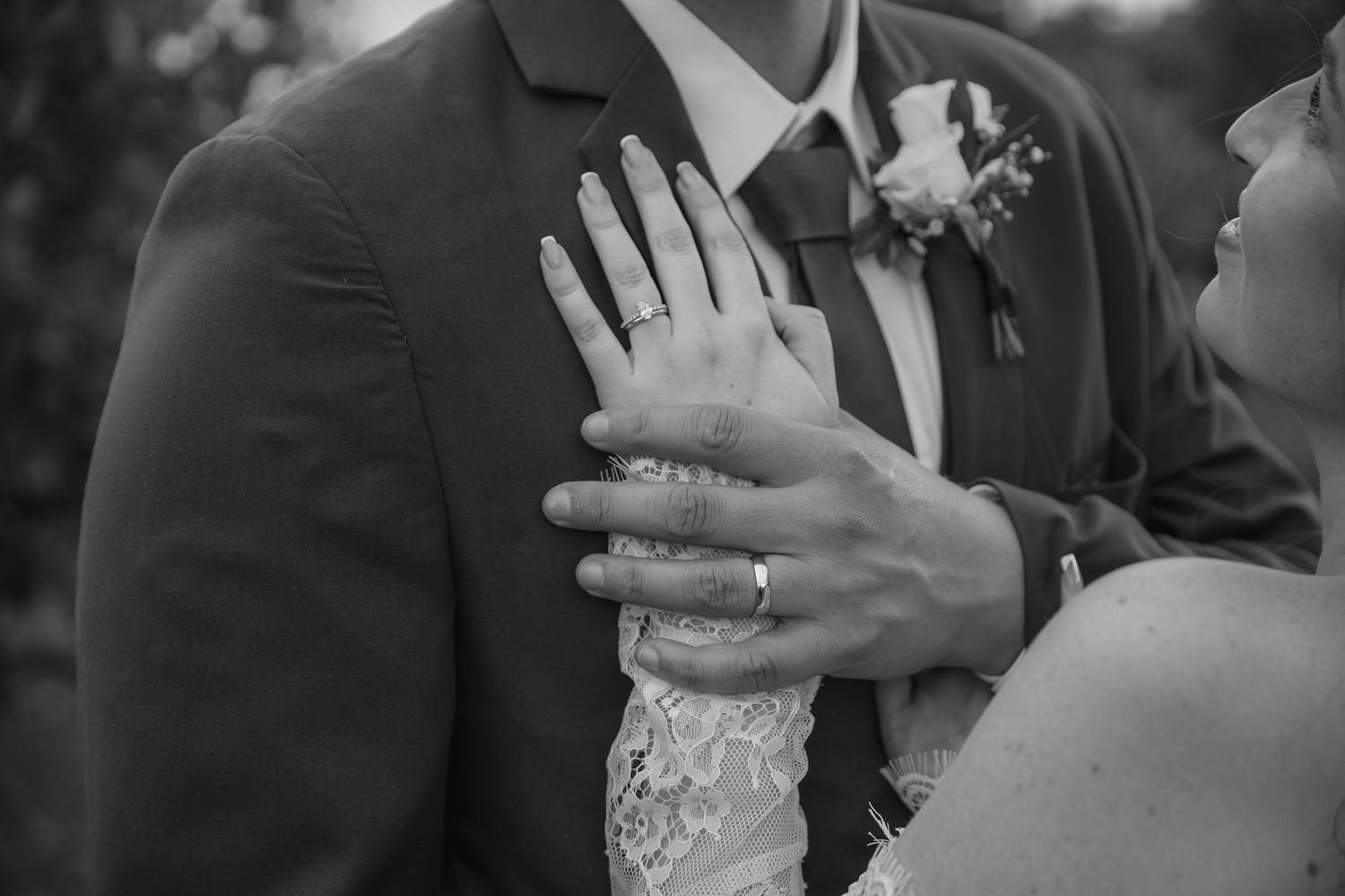 Black and white photo of bride and groom embracing, showcasing wedding rings and lace dress details.