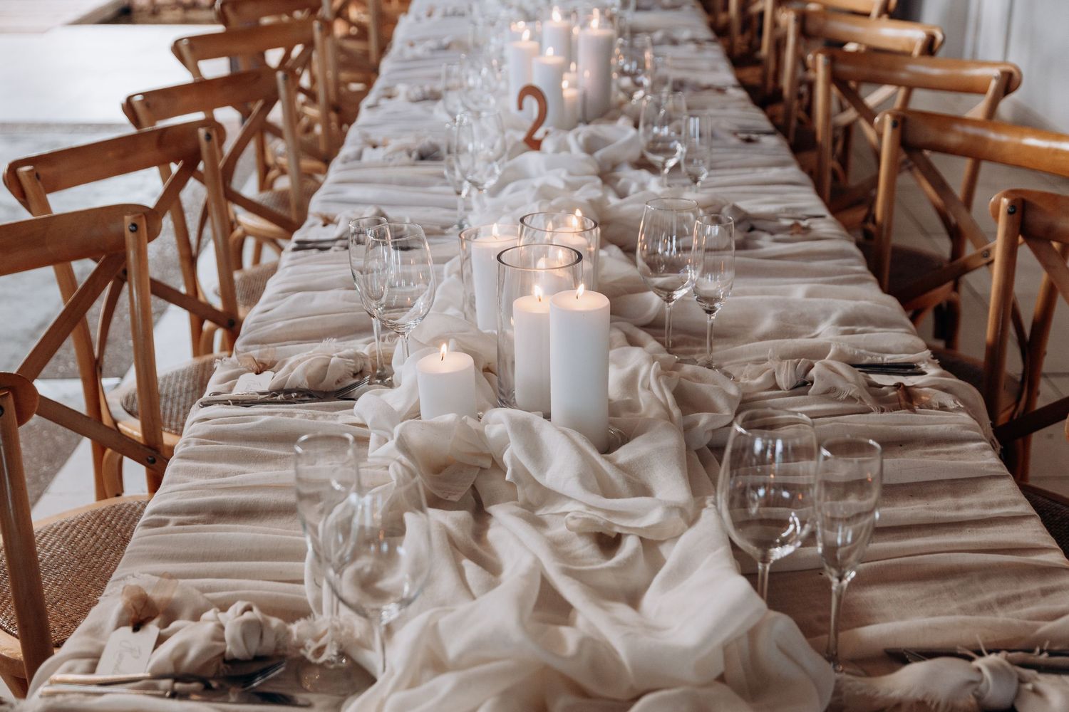 Elegant wedding reception table with white candles, draped linen, crystal glassware, and wooden cross-back chairs.