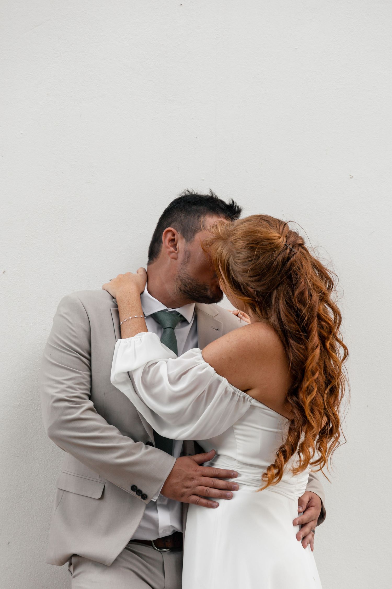A couple in wedding attire embracing and kissing against a white wall, bride in white dress, groom in beige suit.