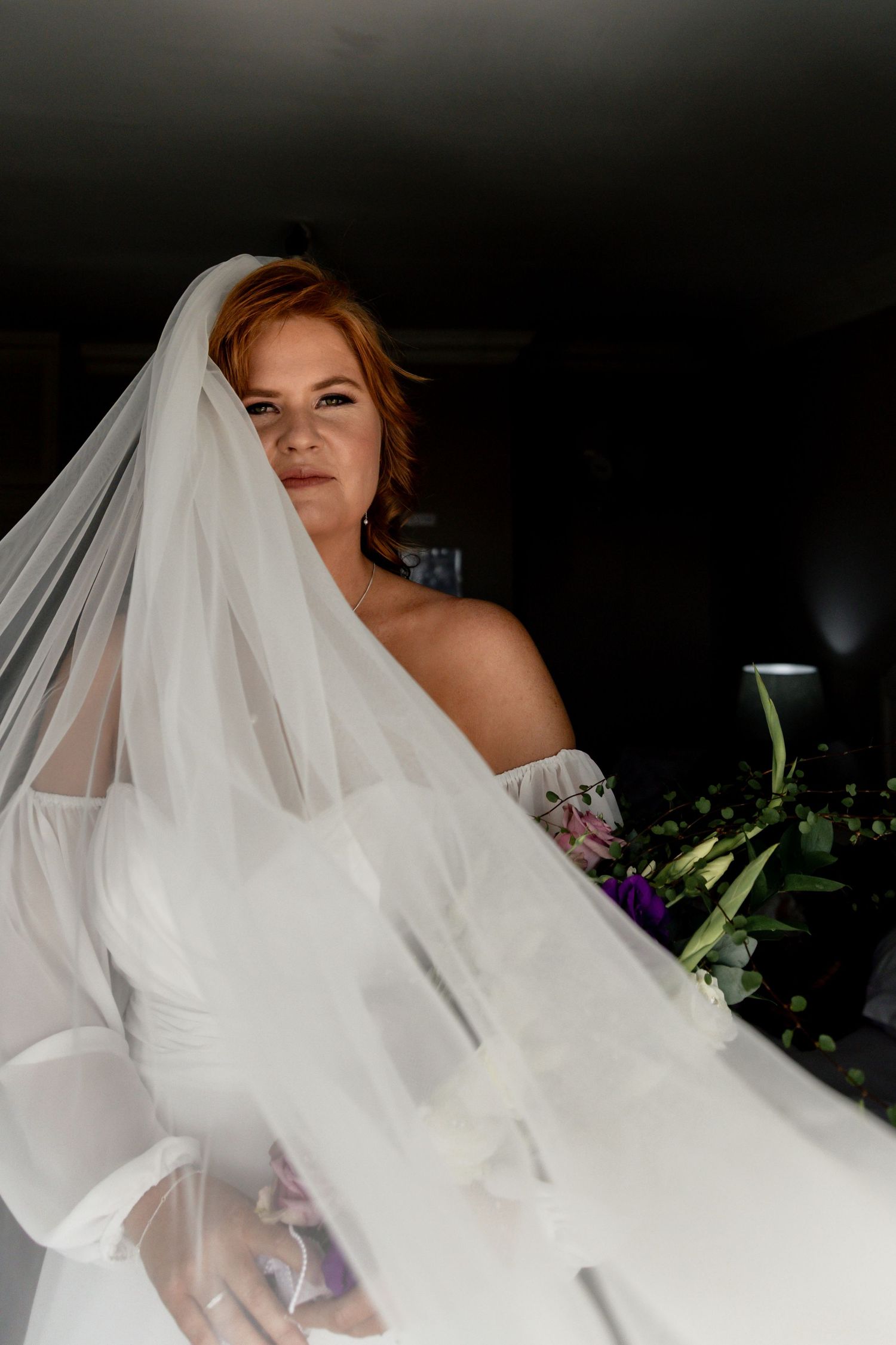Red-haired bride in white off-shoulder gown holding purple bouquet, veil flowing dramatically in soft indoor light.