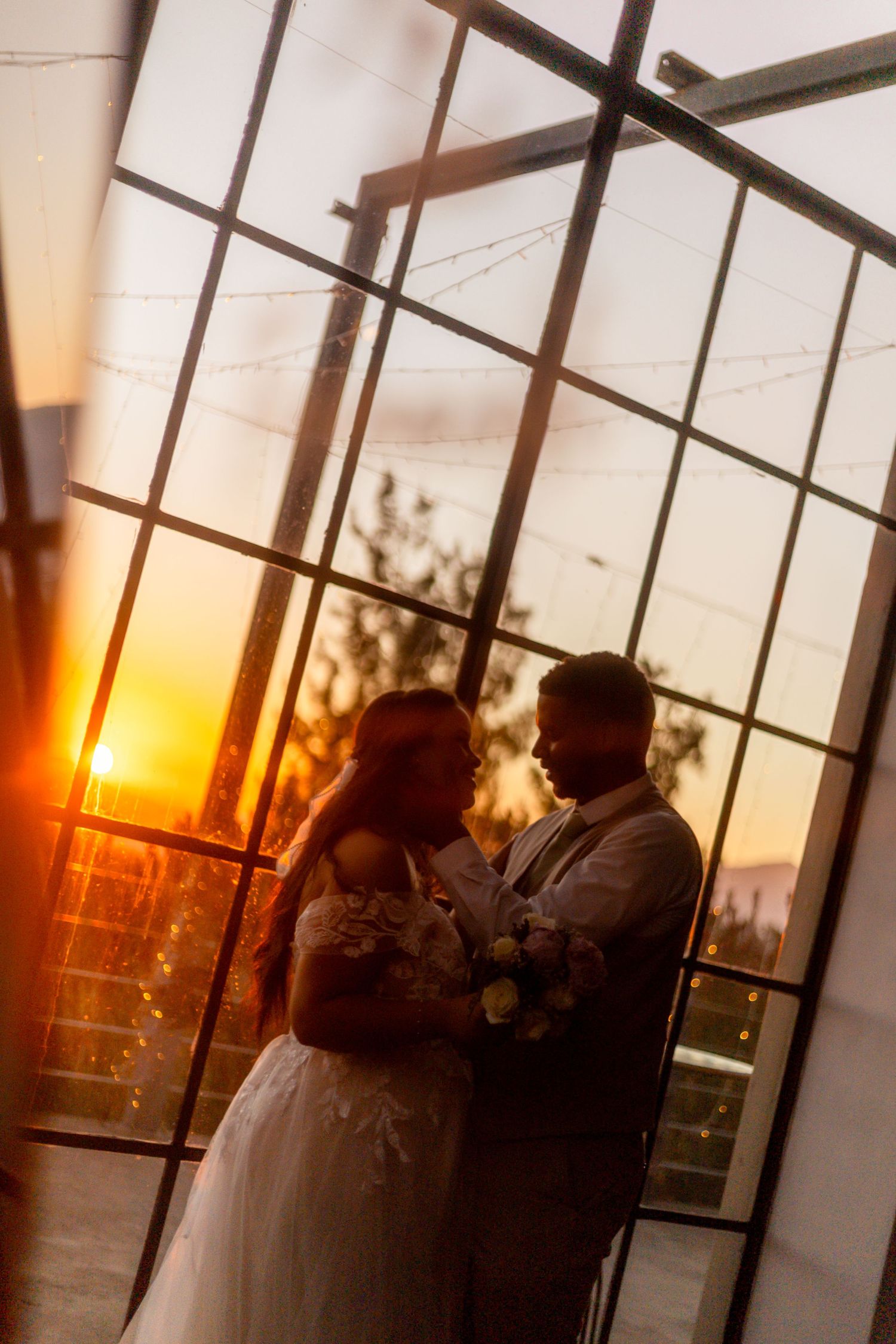 Bride and groom silhouetted against a stunning golden sunset through large glass windows at their wedding venue.