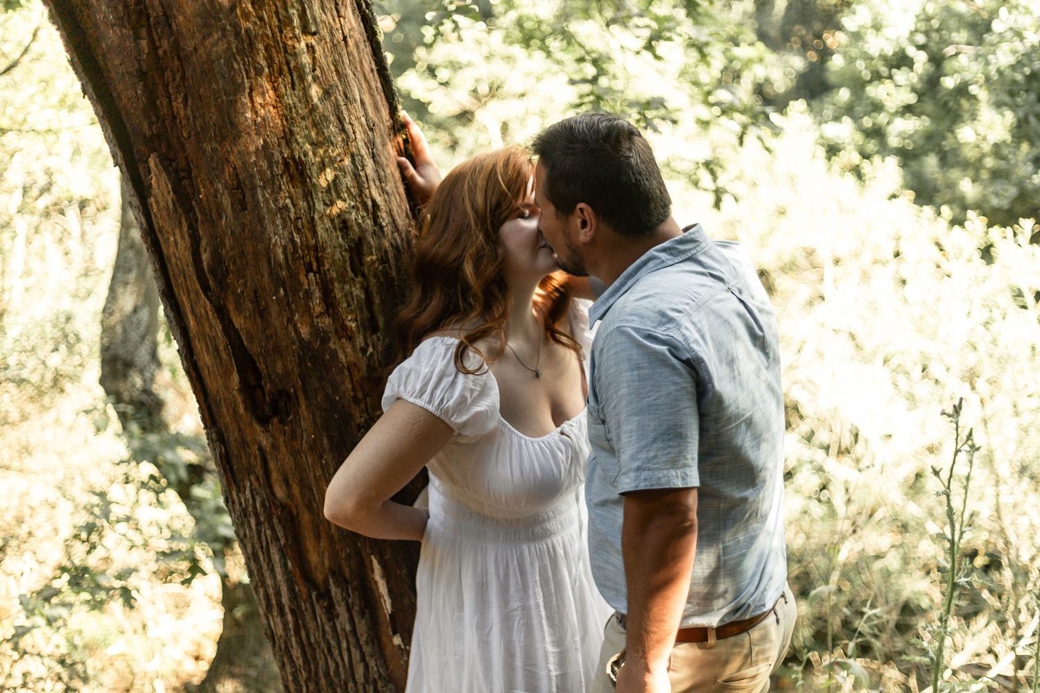 A couple shares a tender moment together in a sunlit forest setting among tall redwood trees.