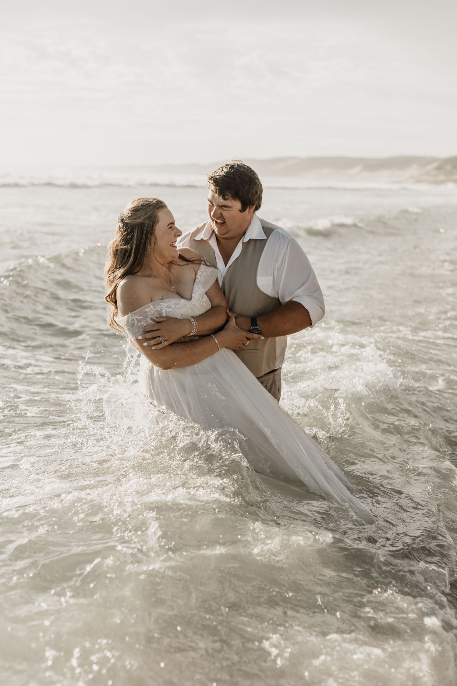 A couple in wedding attire embraces while standing in ocean waves during a romantic beach photoshoot.