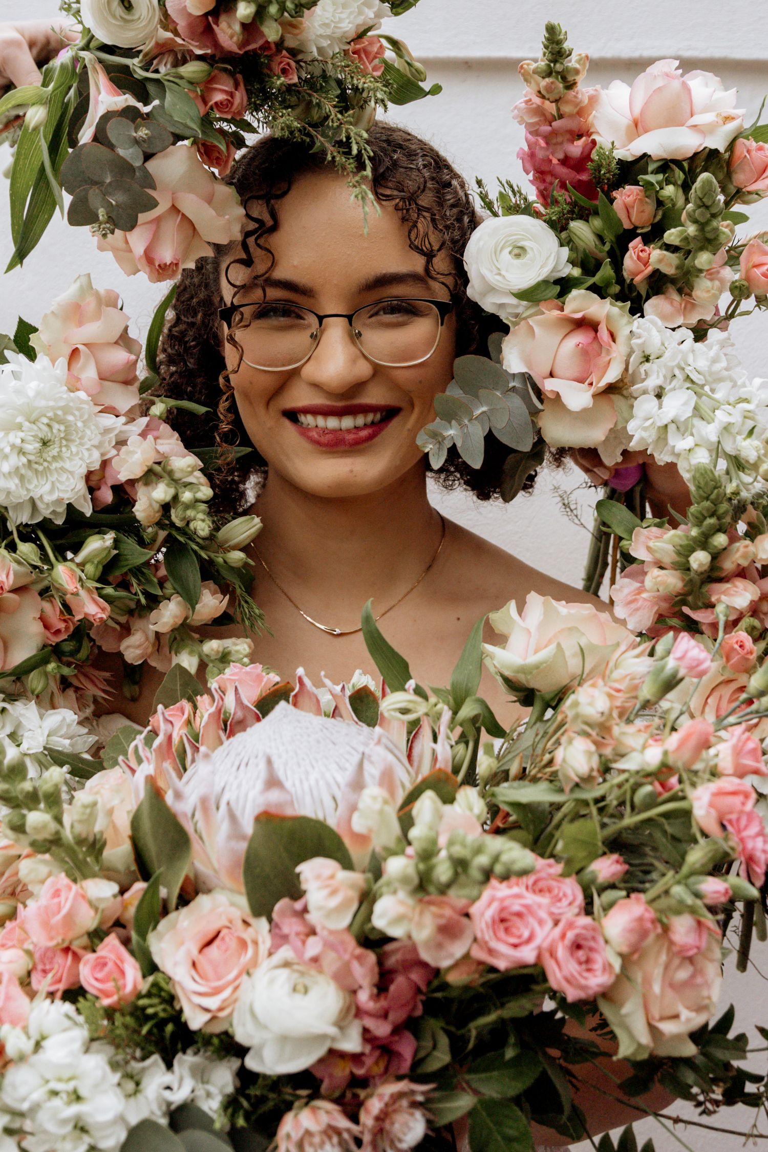 Woman with curly hair and glasses smiling amid a lush floral arrangement of pink and white roses with greenery.