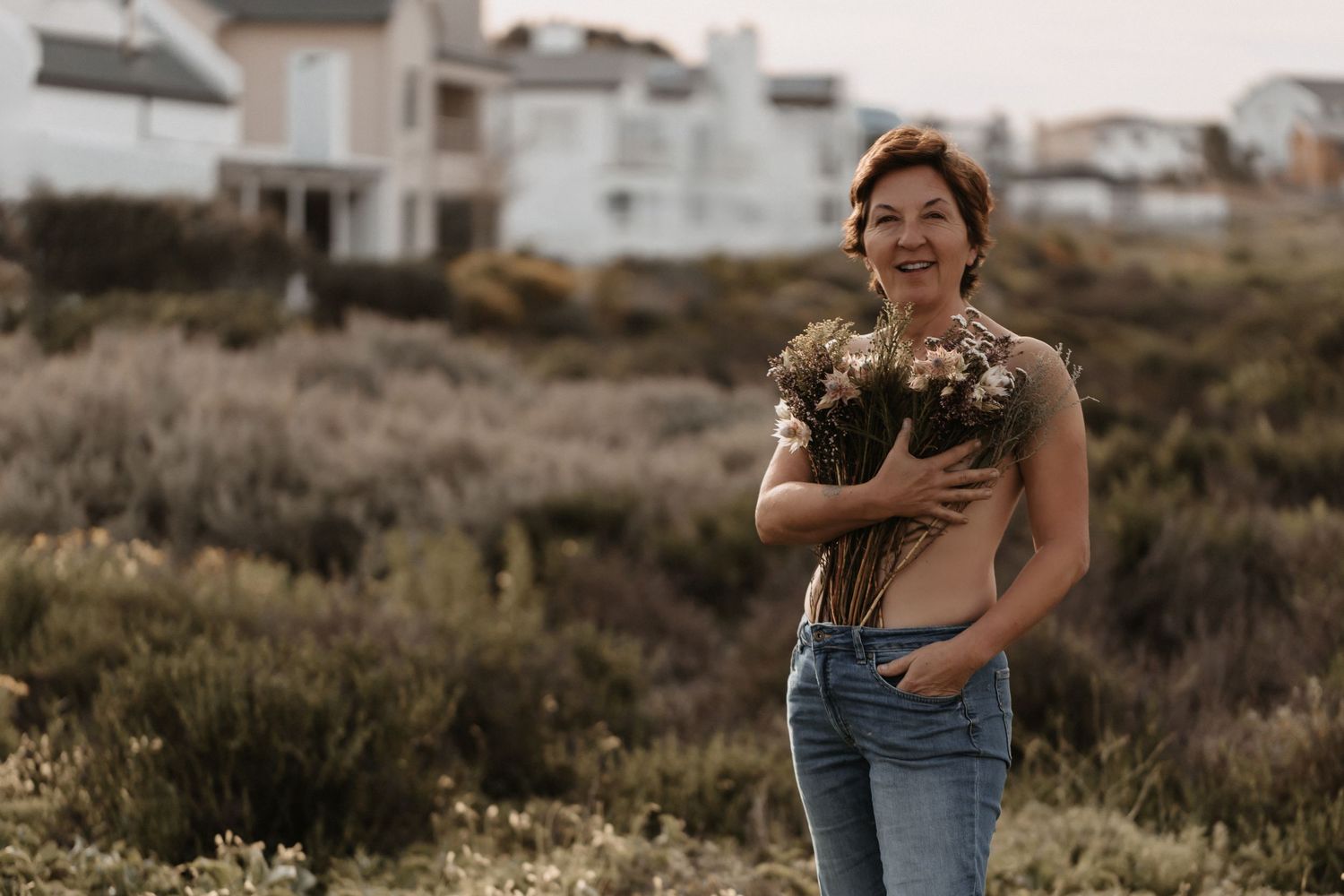Someone wearing blue jeans and holding a bouquet of flowers stands in a field with coastal houses in the background.