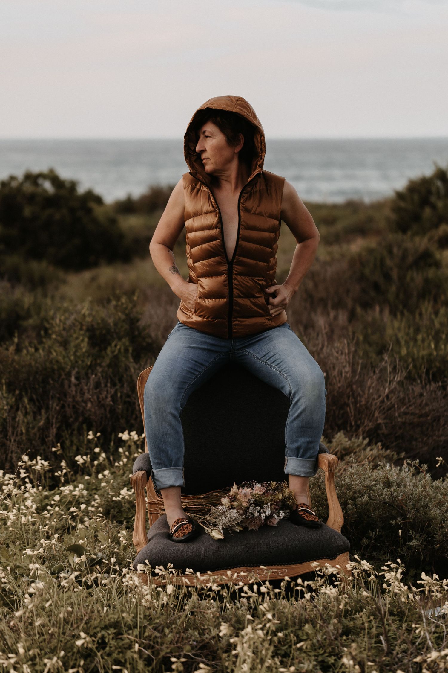 Someone in a brown puffer vest and jeans sits on a chair in a coastal meadow at sunset with wildflowers blooming.