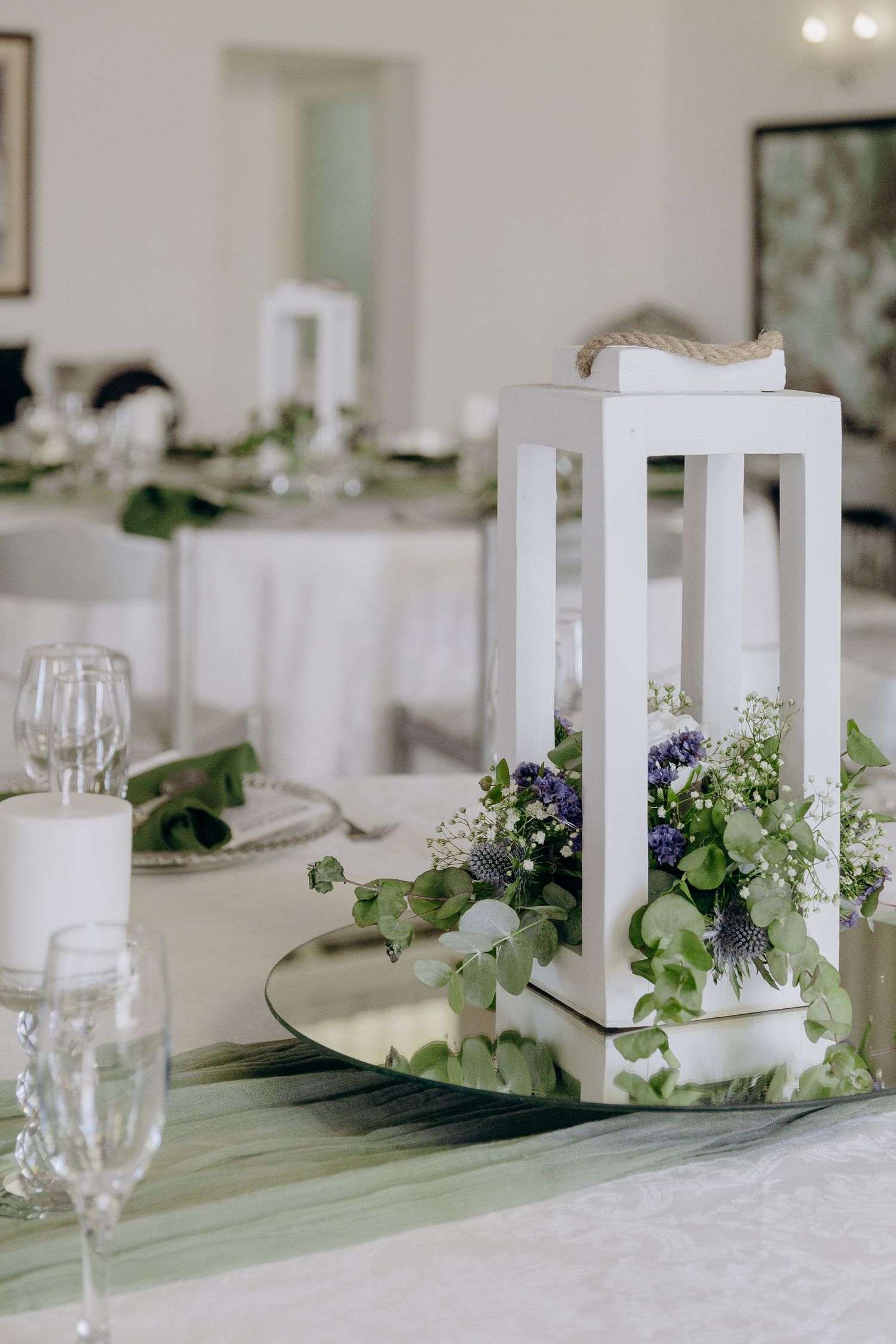 Elegant white lantern centerpiece with greenery and purple flowers on a wedding reception table with crystal glassware.
