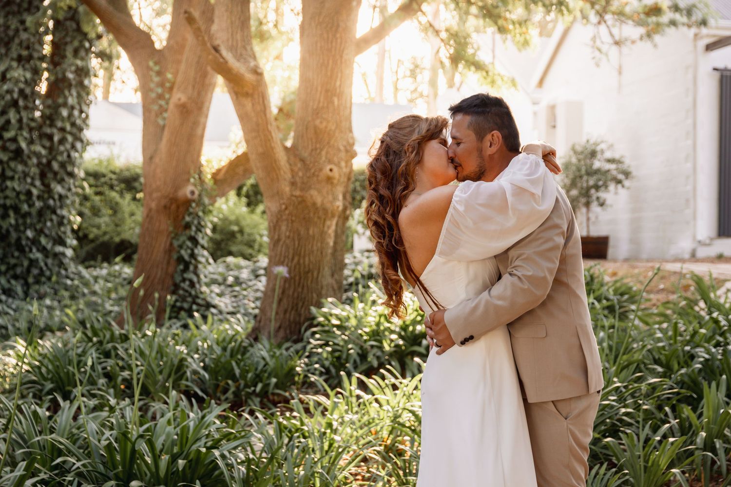 Bride and groom sharing a romantic kiss in a sunlit garden, surrounded by lush green foliage.