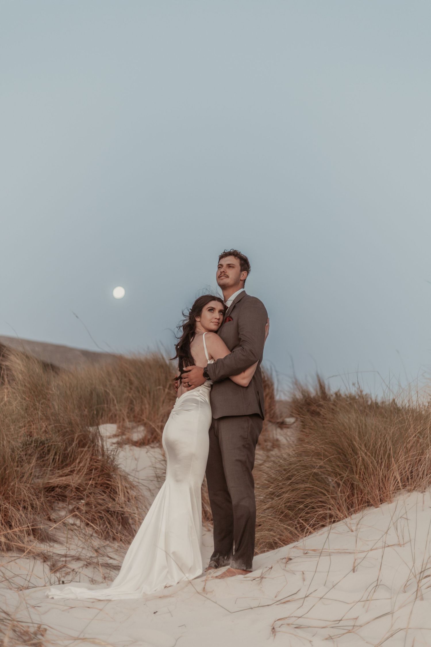 Bride and groom embracing on sand dunes under a full moon, surrounded by coastal grass at twilight.