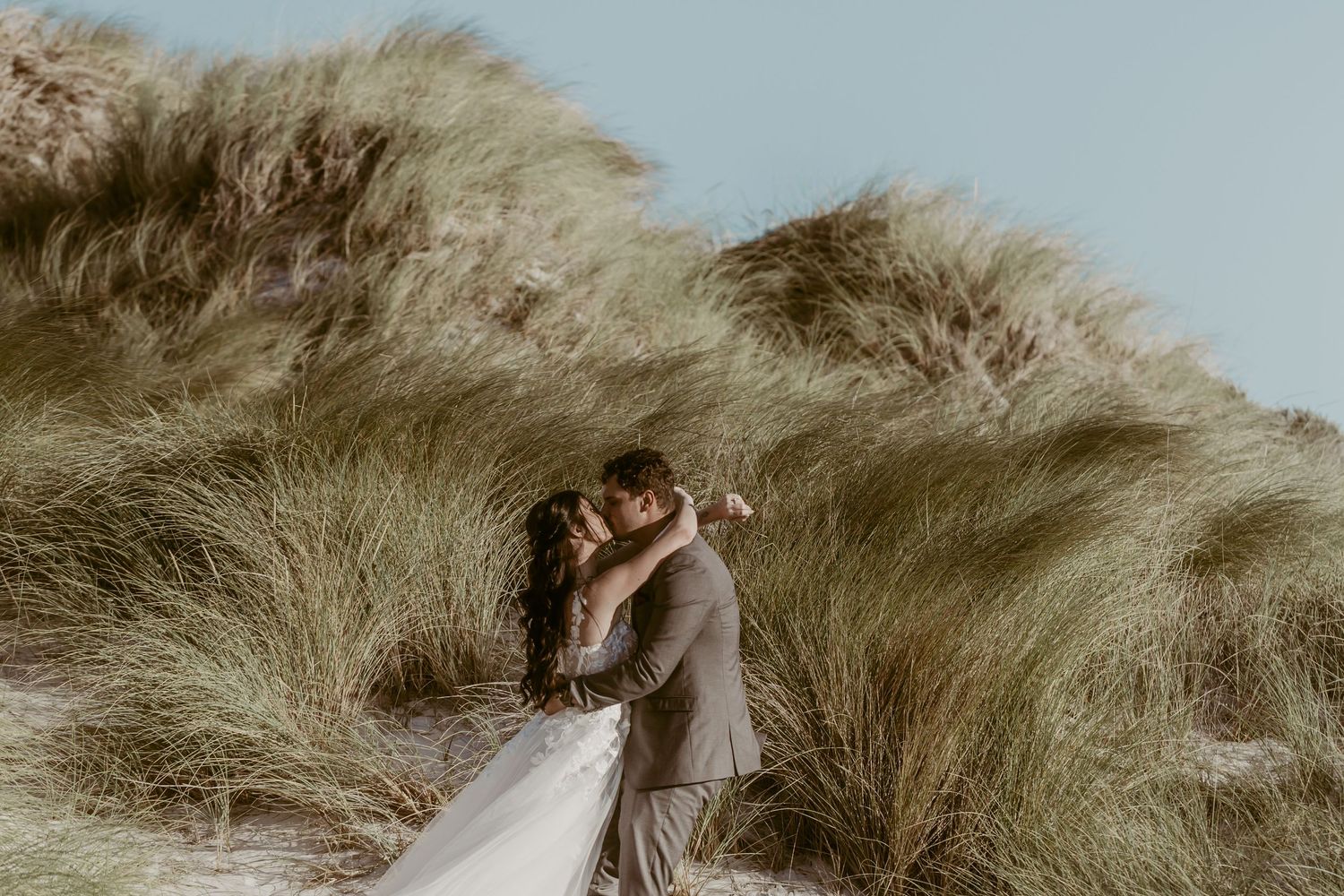 Bride and groom sharing a romantic kiss among sandy dunes with tall green grass under a clear blue sky.