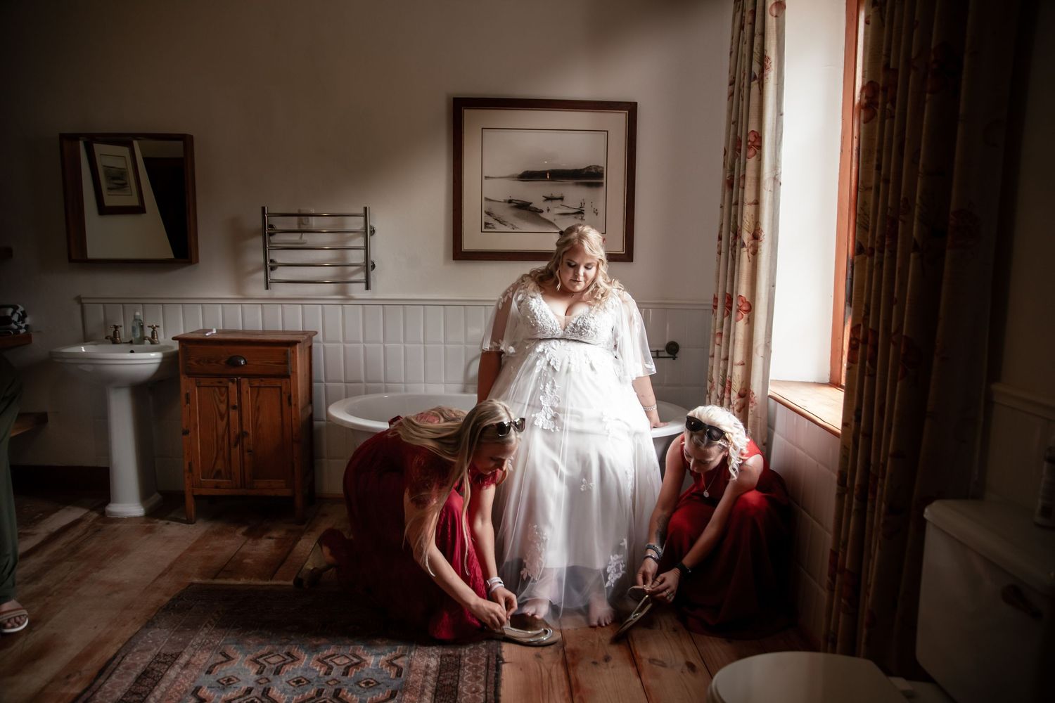 Bride in white wedding dress being helped with shoes by two bridesmaids in red dresses in a vintage bathroom.